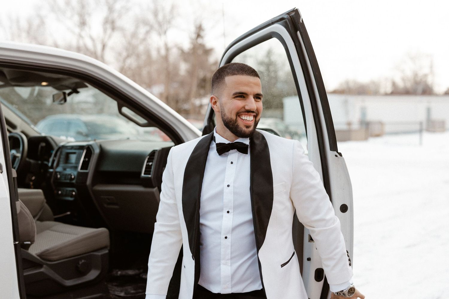 Groom in white tuxedo jacket and bowtie steps out of car on snowy winter day.