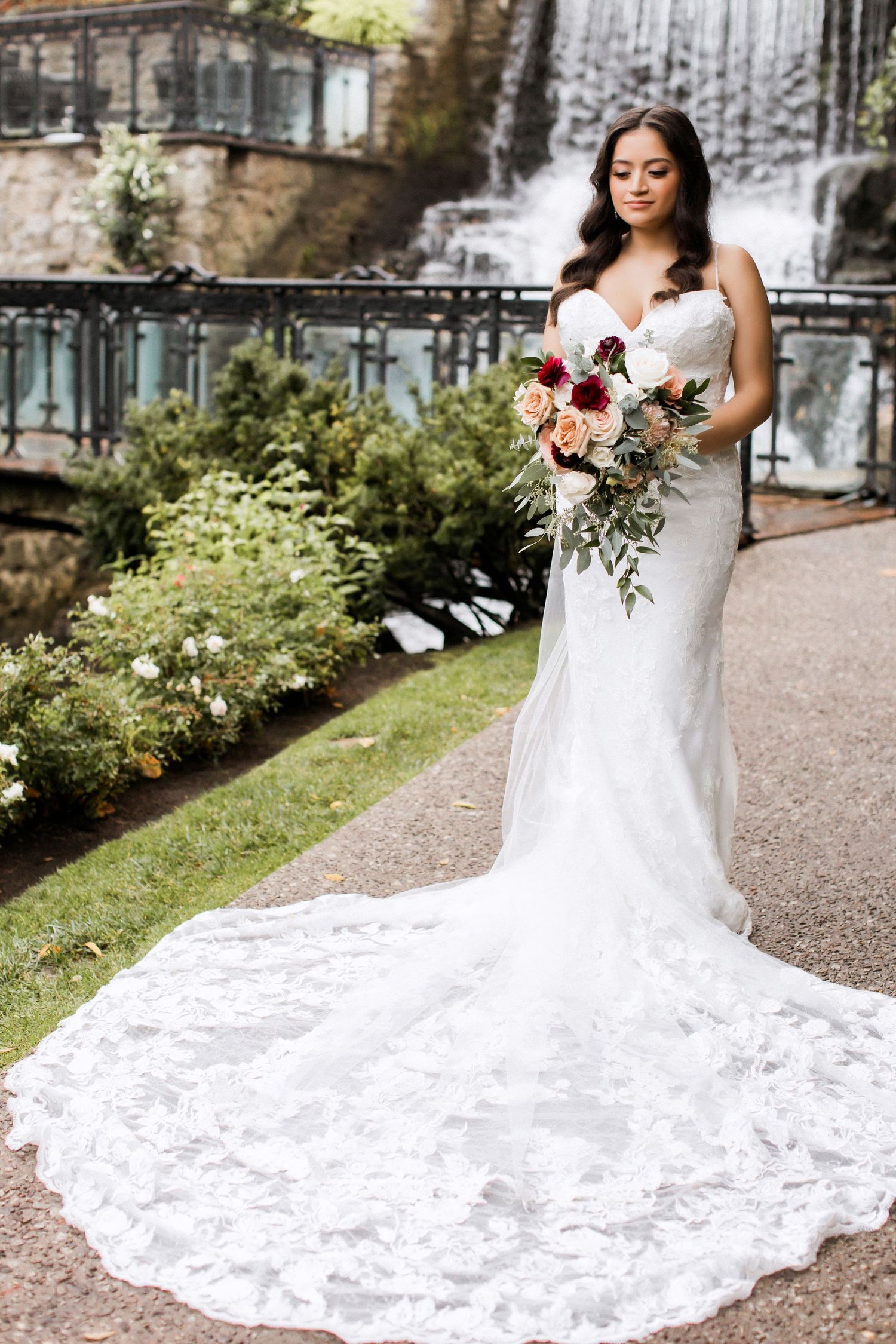 Bride in fitted lace gown holds bouquet while posing in front of waterfall.