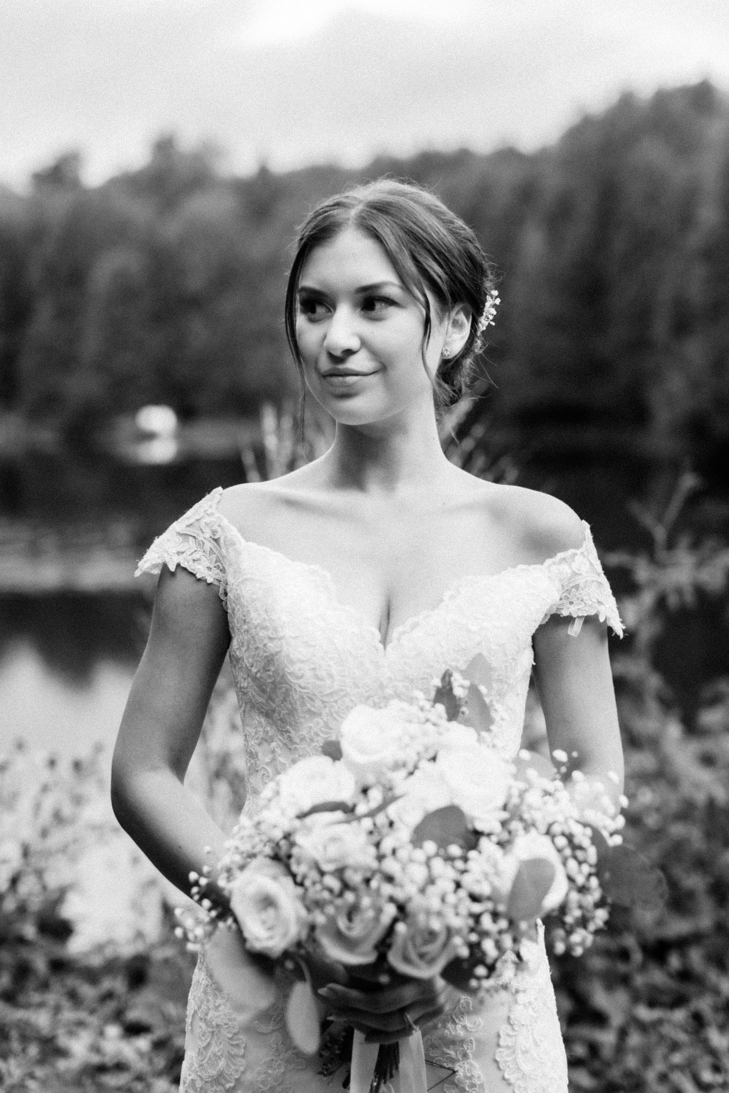 Elegant bride in lace cap sleeve dress holds white bouquet by lakeside.