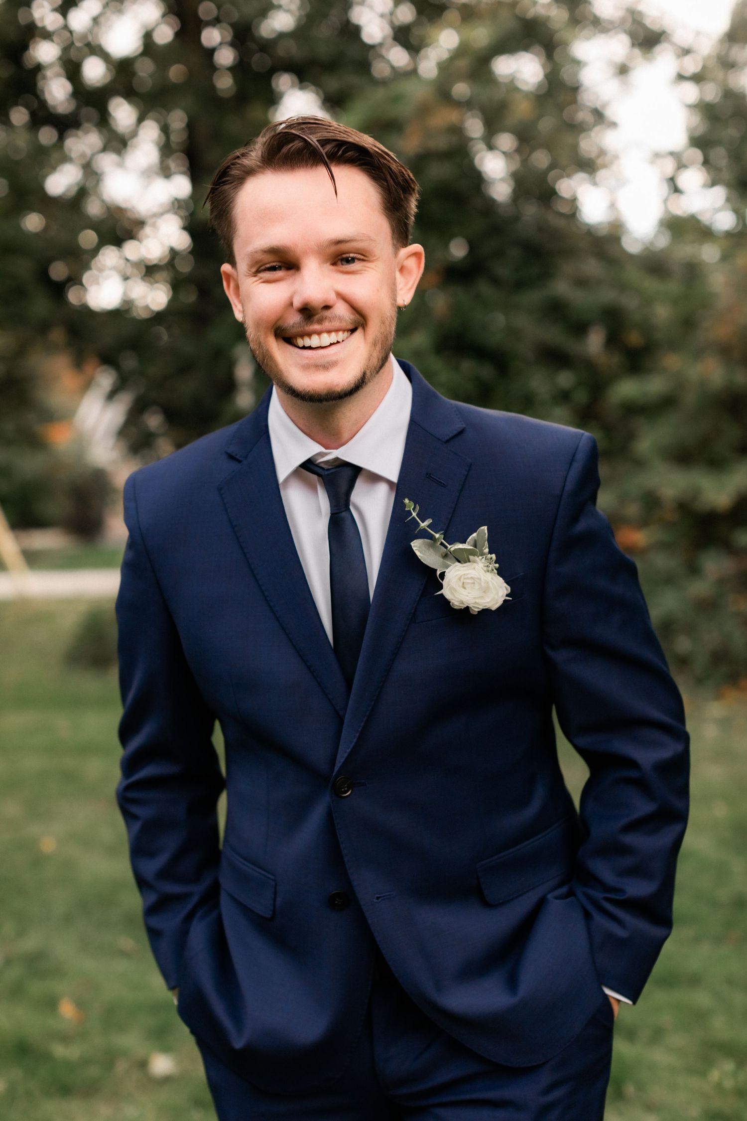 Groom in navy blue suit with boutonniere smiling outdoors in garden setting.