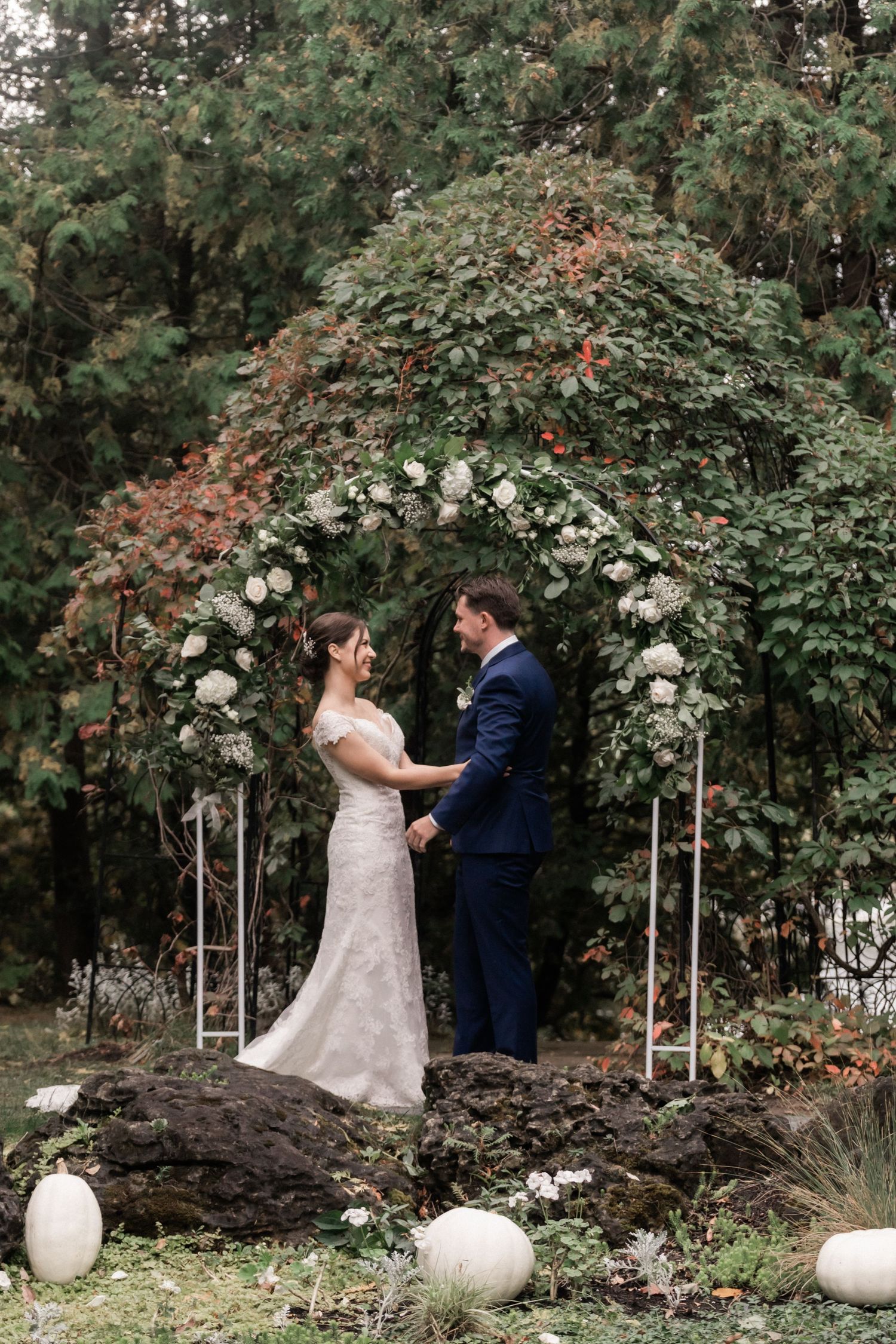 Wedding couple stands under floral arch with white pumpkins decorating the autumn garden setting.