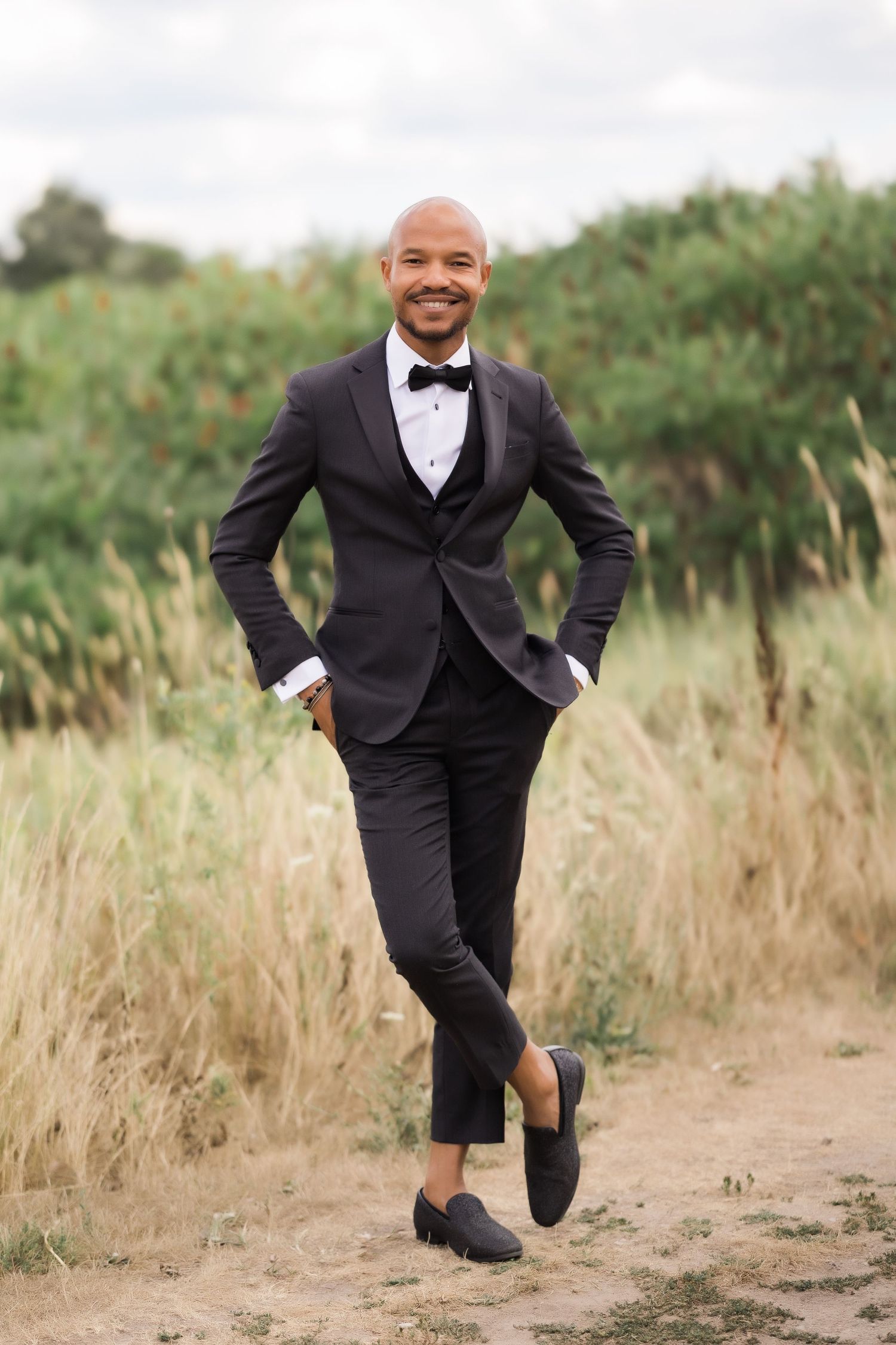 Smiling groom shows off stylish tuxedo while posing casually in tall grass.