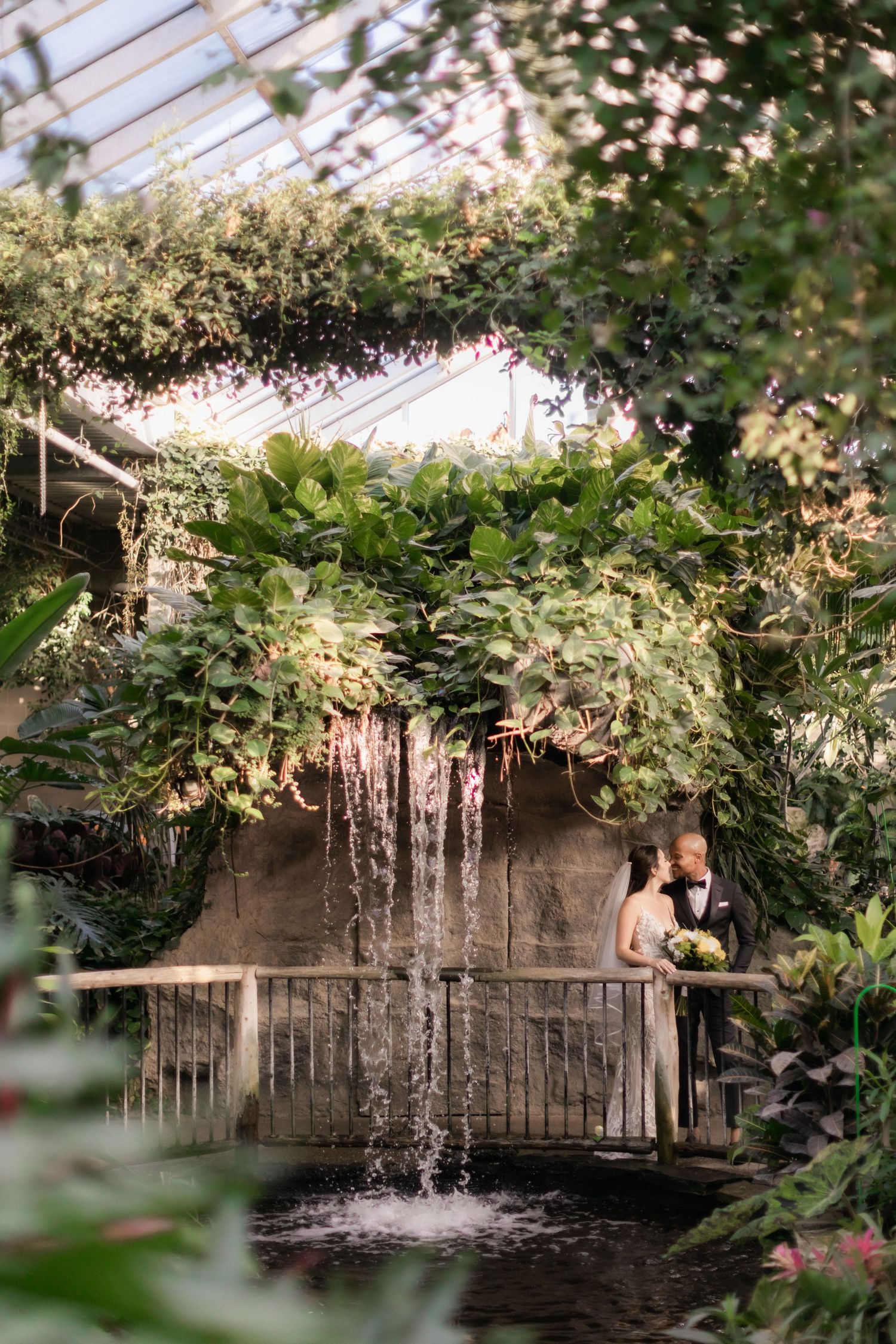 Wedding couple stands by cascading waterfall in greenhouse surrounded by lush foliage.