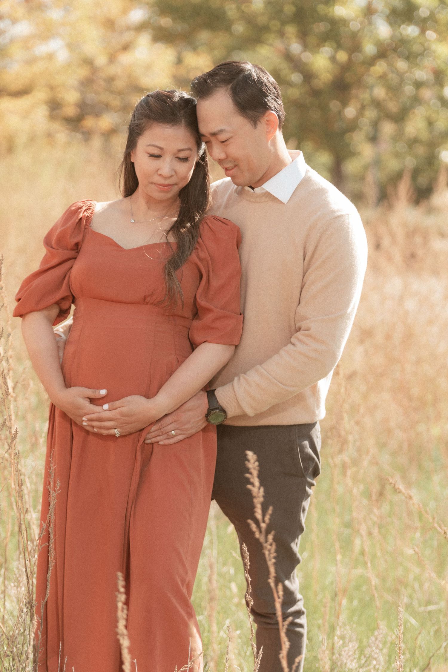 Tender moment between expectant parents standing among tall autumn grasses at sunset.