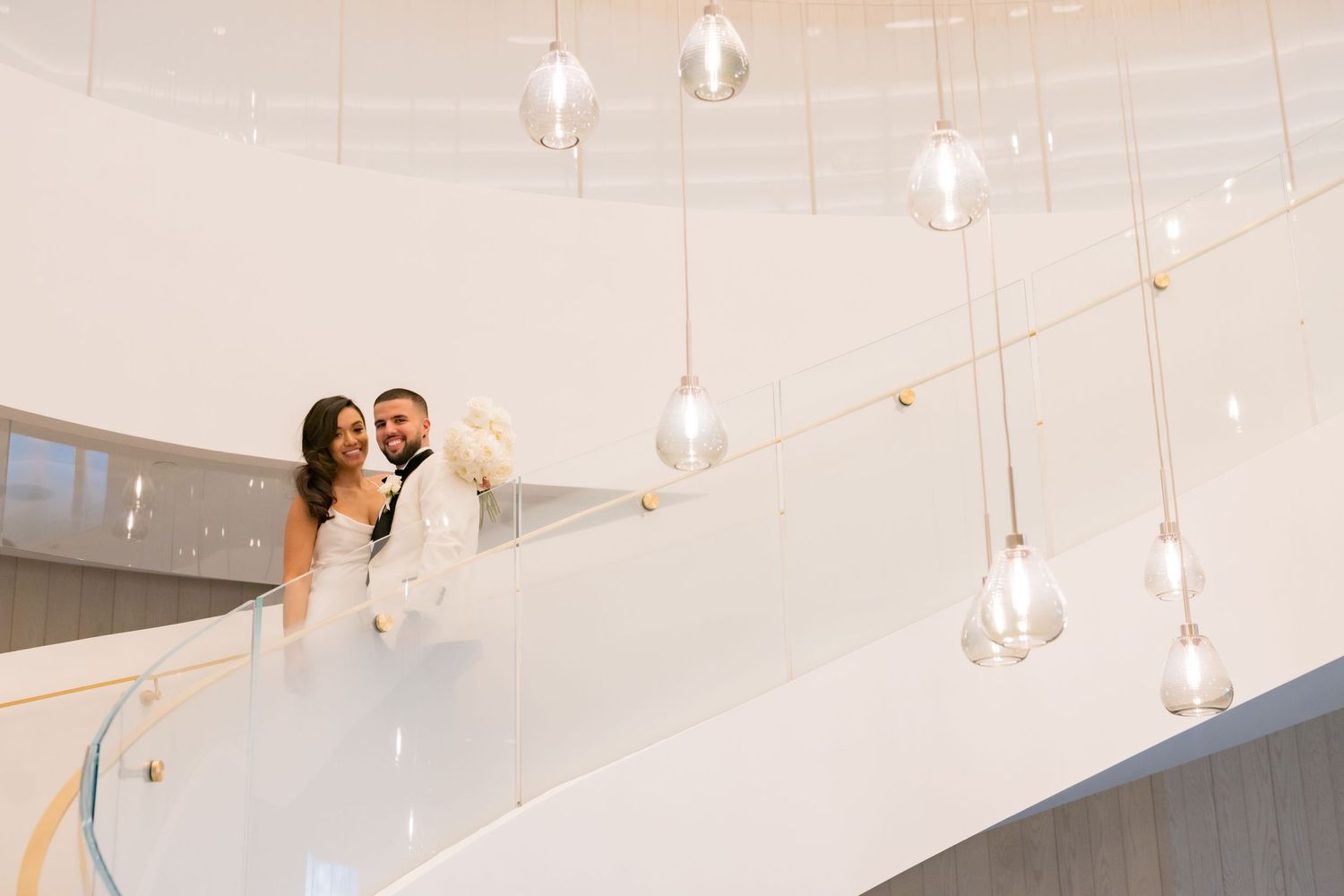 Modern wedding portrait on white spiral staircase with suspended glass light fixtures.