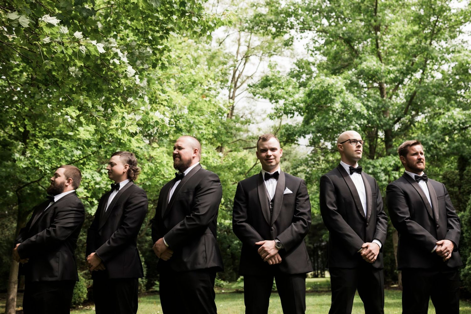 Wedding party in formal wear arranged in a line outdoors for photos among garden greenery.