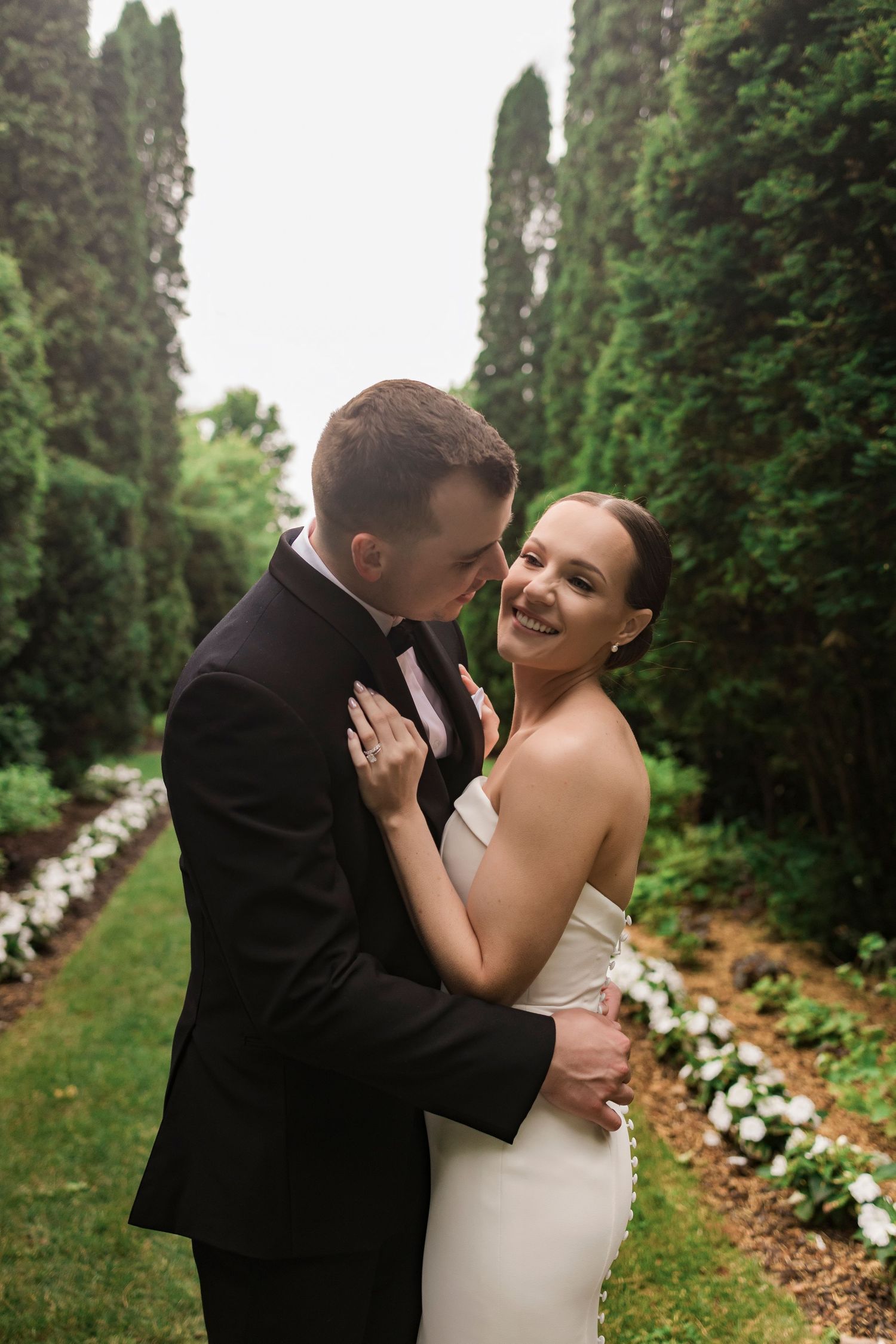 Romantic wedding portrait of a couple embracing in a garden path lined with tall cypress trees and flowers.