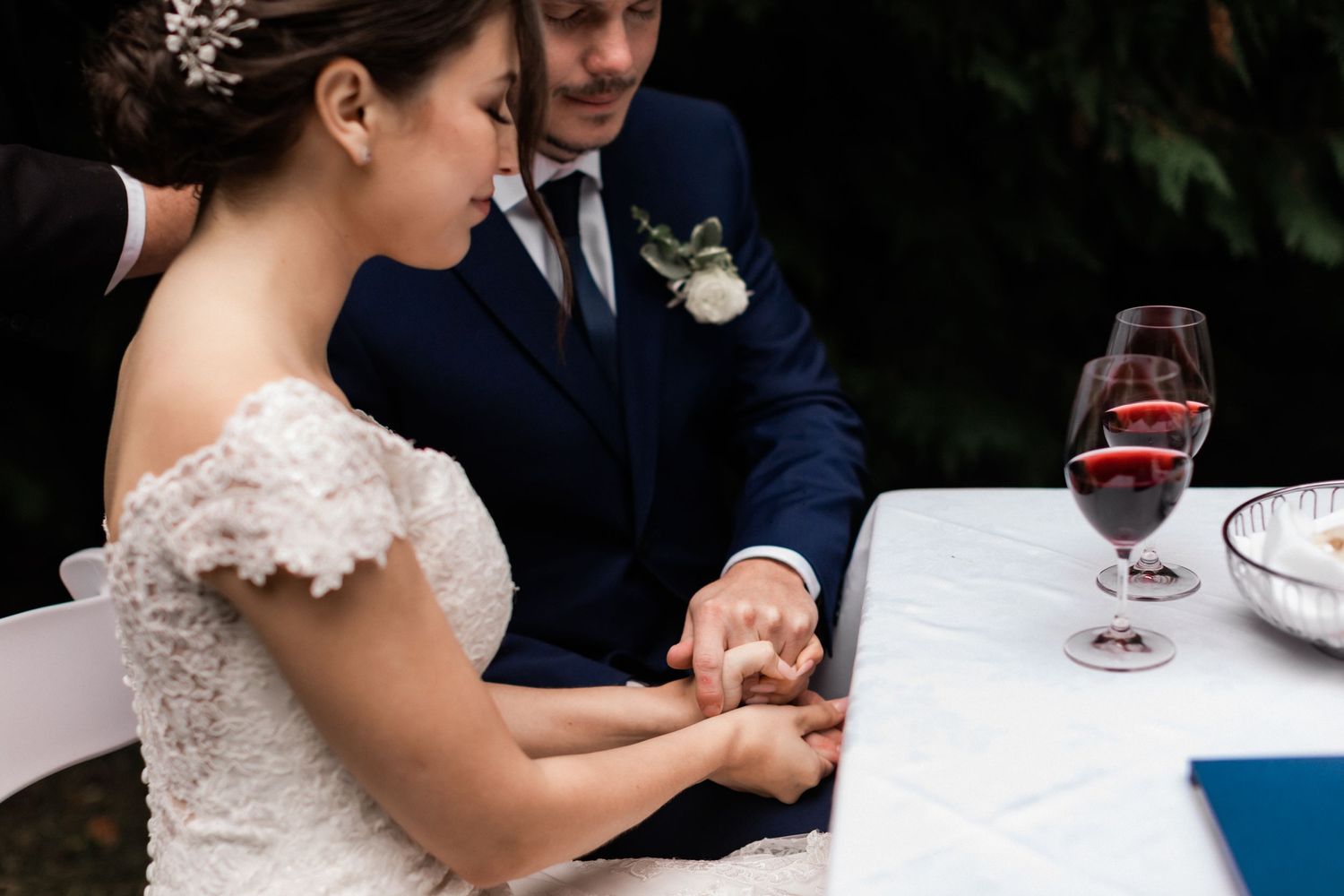 Close-up of couple's hands during intimate outdoor wedding ceremony with wine glasses on table.