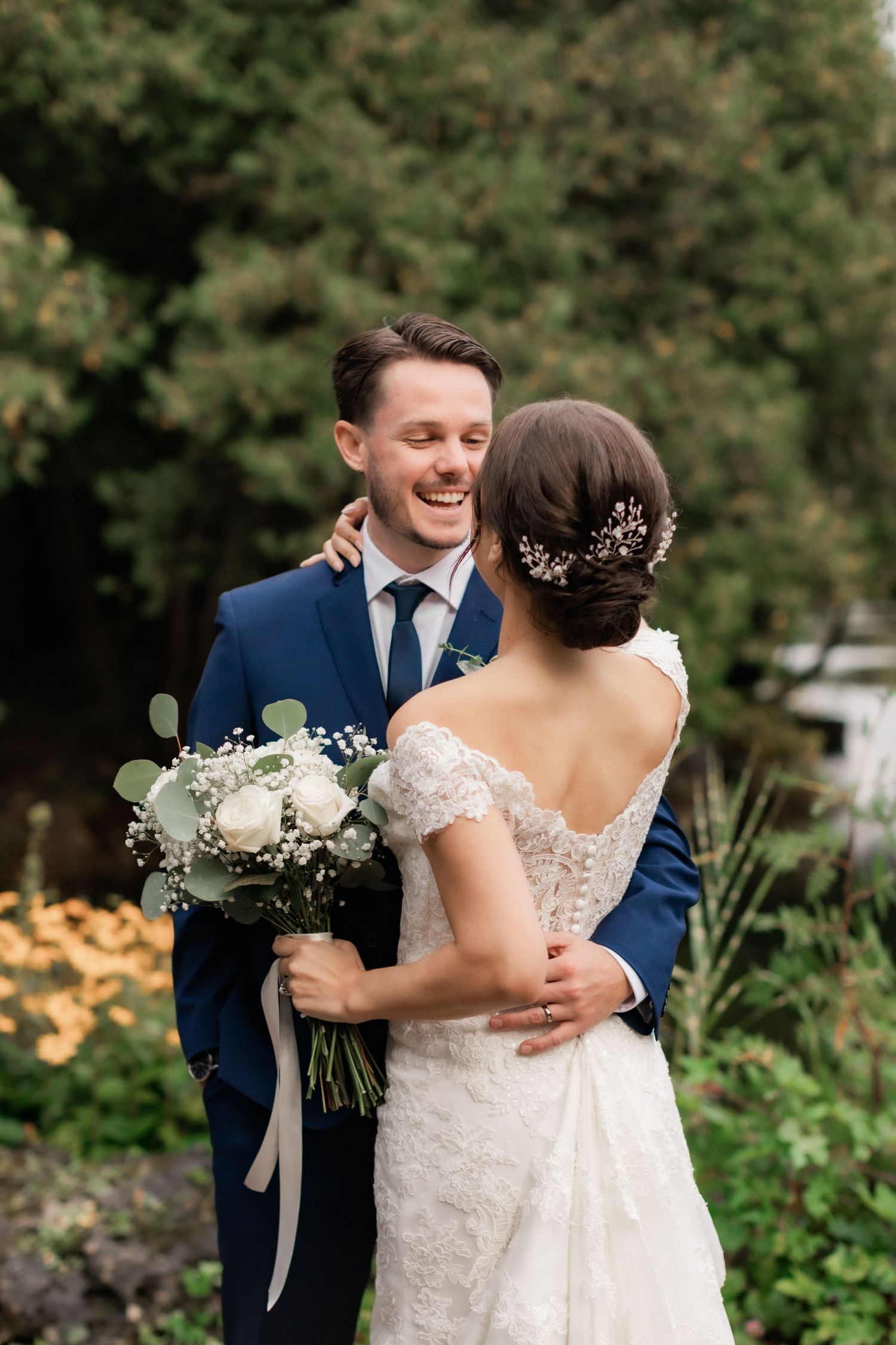 Newlyweds share a joyful moment outdoors with a white floral bouquet against a natural garden backdrop.