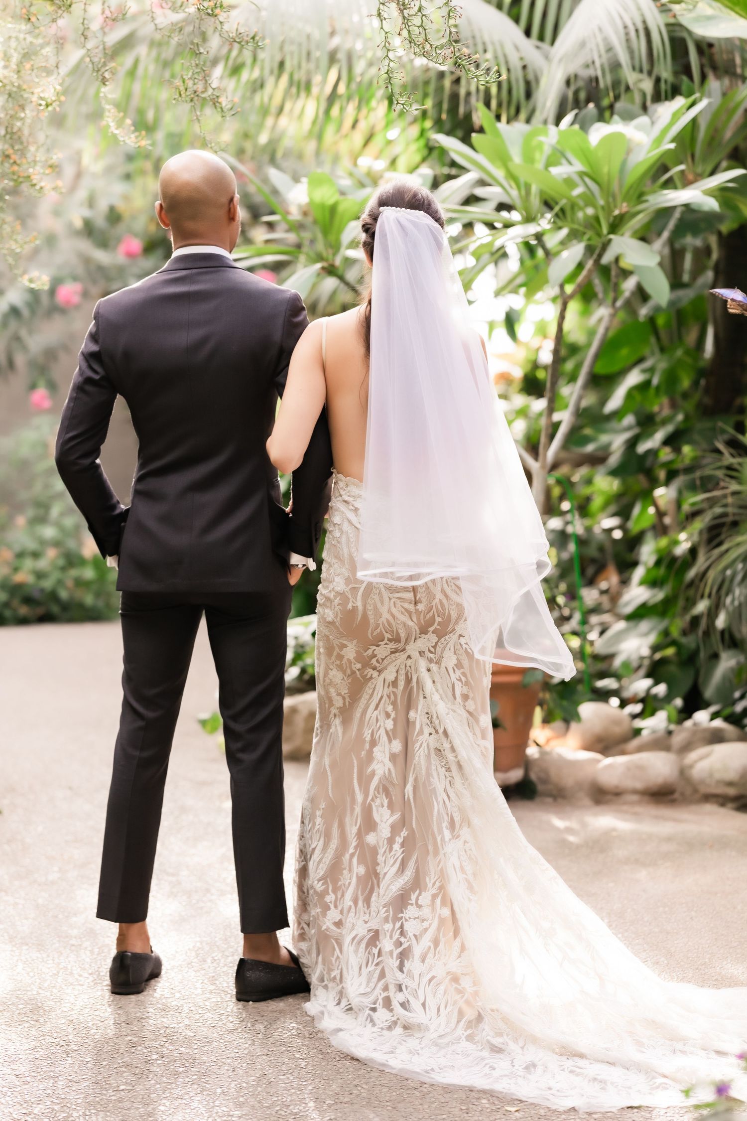 Back view of newlyweds showing detailed lace wedding dress and classic tuxedo in garden setting.