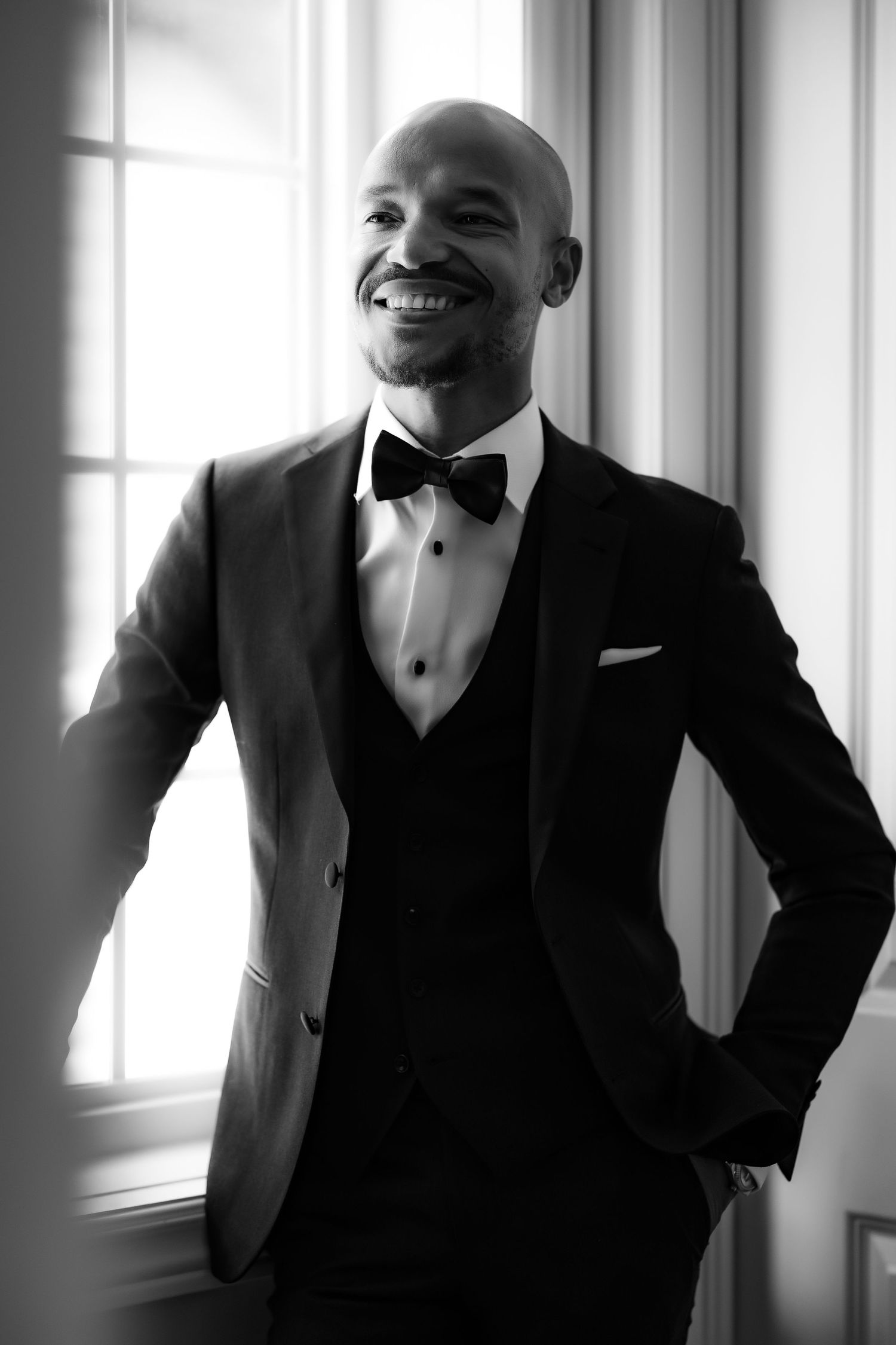 Black and white portrait of groom in fitted tuxedo standing by window with natural light.