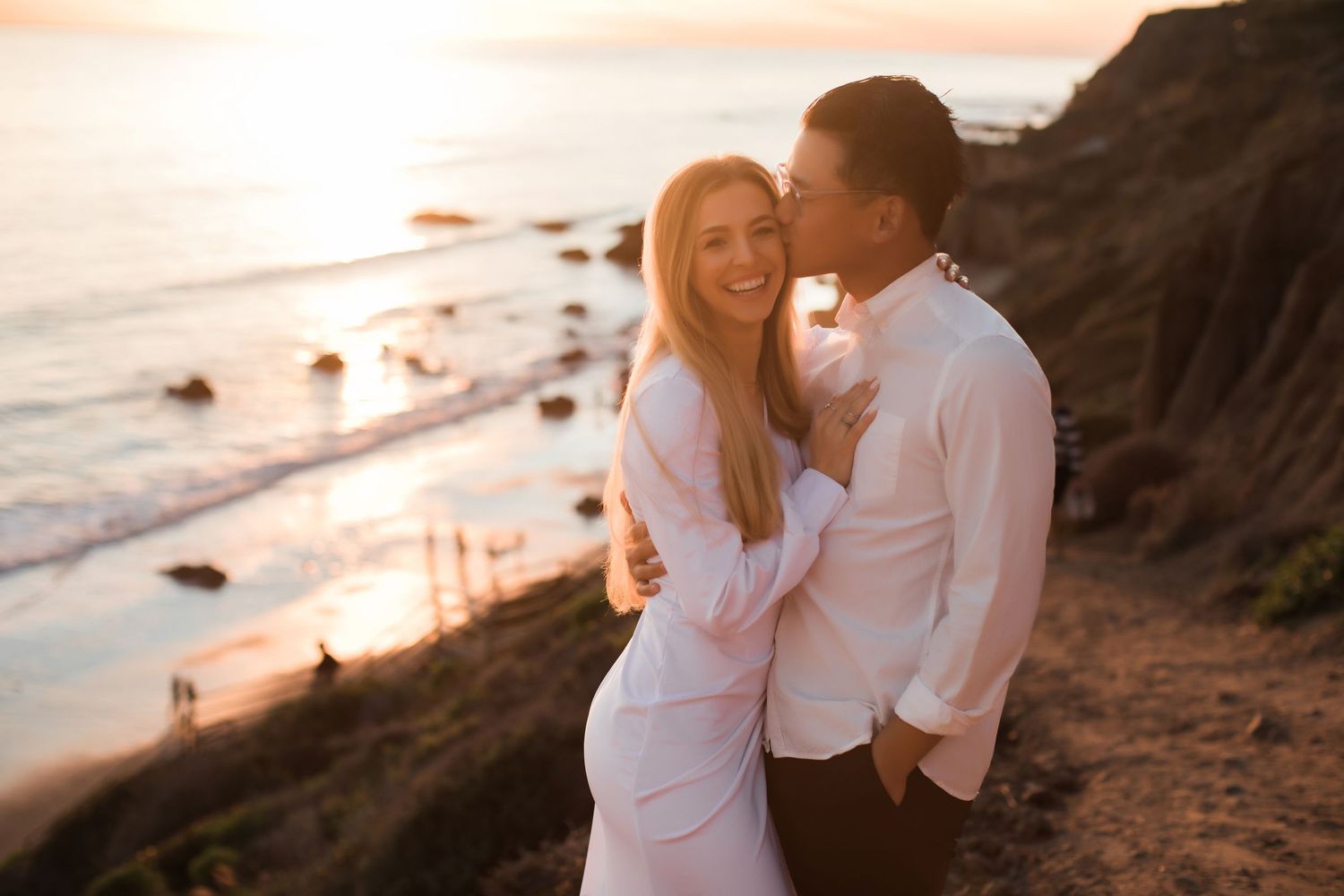 Couple shares intimate moment on rocky coastline at sunset wearing matching white outfits.
