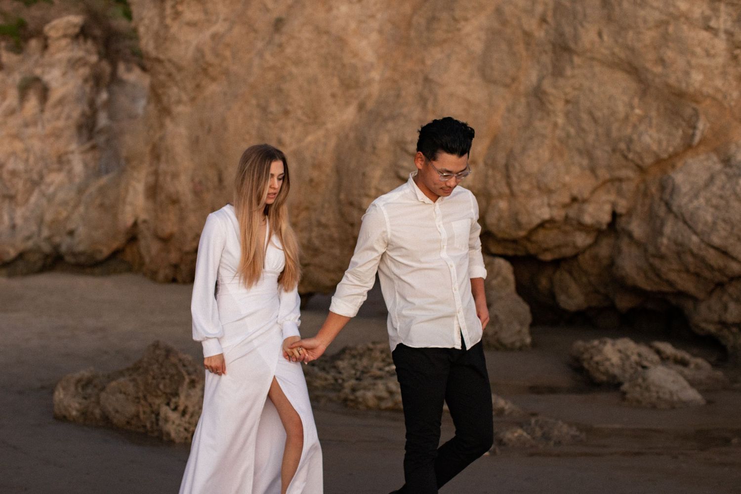 Couple walking hand in hand along rocky beach at sunset.