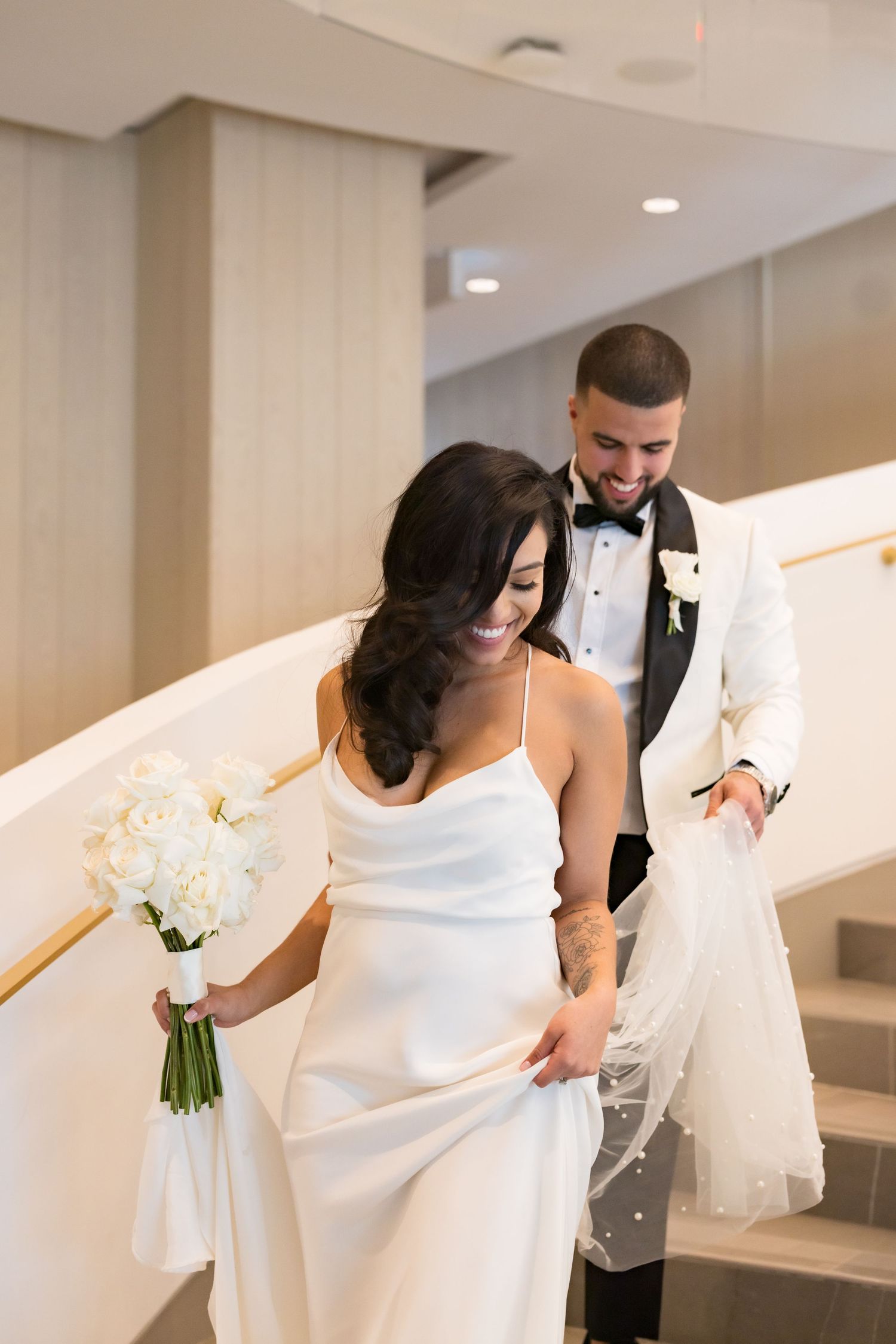 Wedding couple descending white curved staircase with bride holding bouquet of white roses.