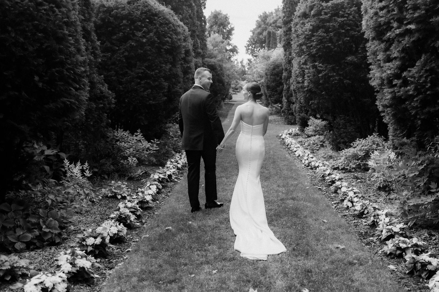 Black and white photo of couple walking down garden path lined with flowers and hedges.