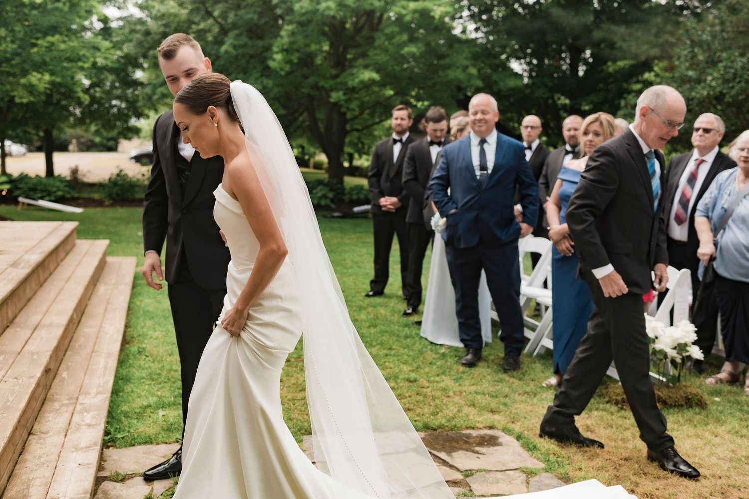 Joyful wedding couple holds hands and laughs while standing on wooden steps during outdoor garden ceremony.