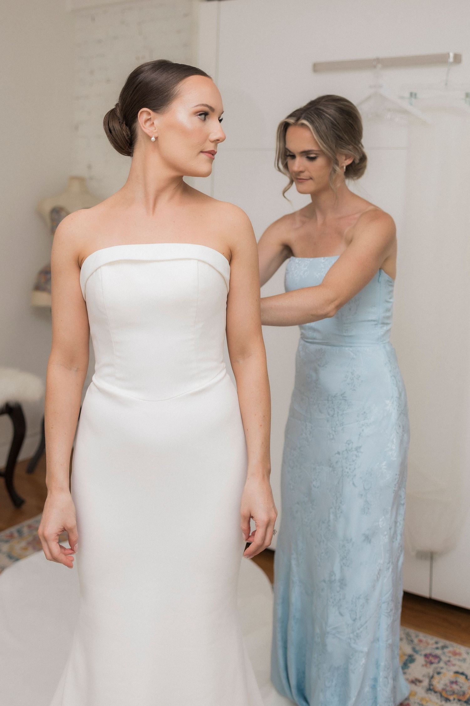 A bride in a strapless white wedding gown stands while an attendant in a light blue dress helps with preparations.