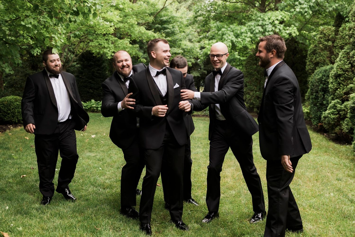 A group dressed in formal black tuxedos gather outdoors sharing a lighthearted moment on a grassy lawn.