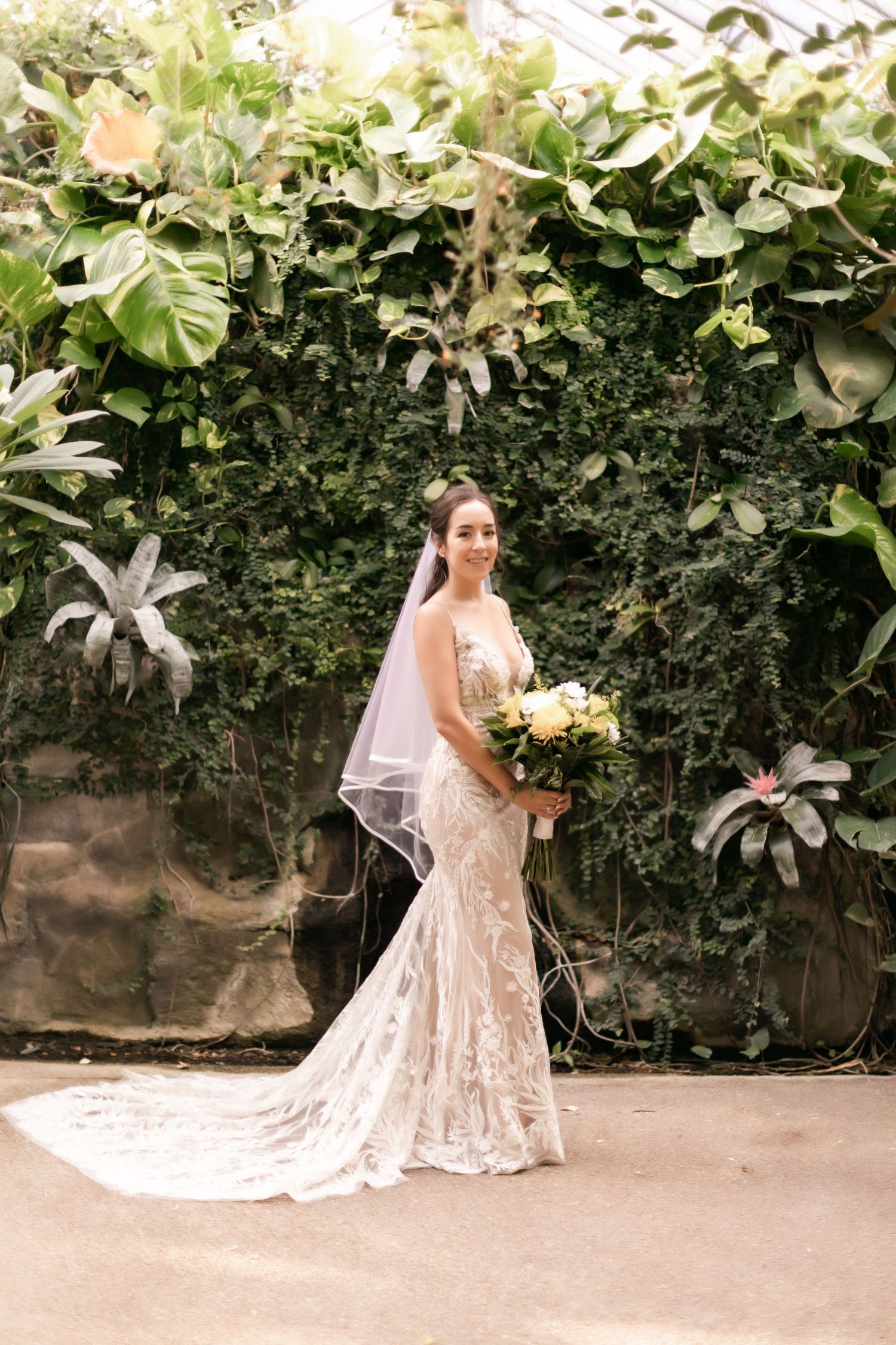 Bride in lace mermaid gown with long veil poses with bouquet against garden backdrop.