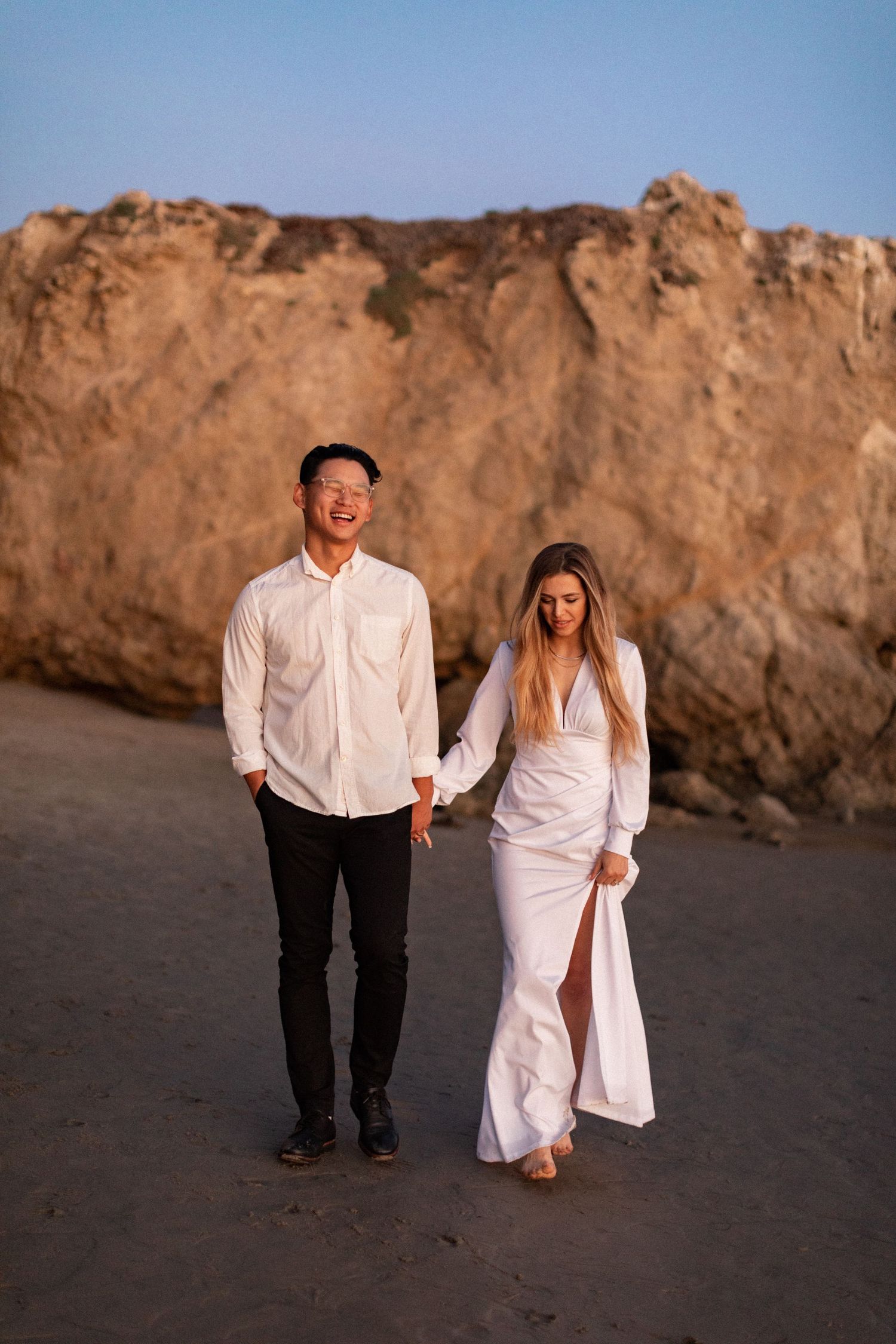 Happy couple walking together near beach cliffs during golden hour photoshoot.