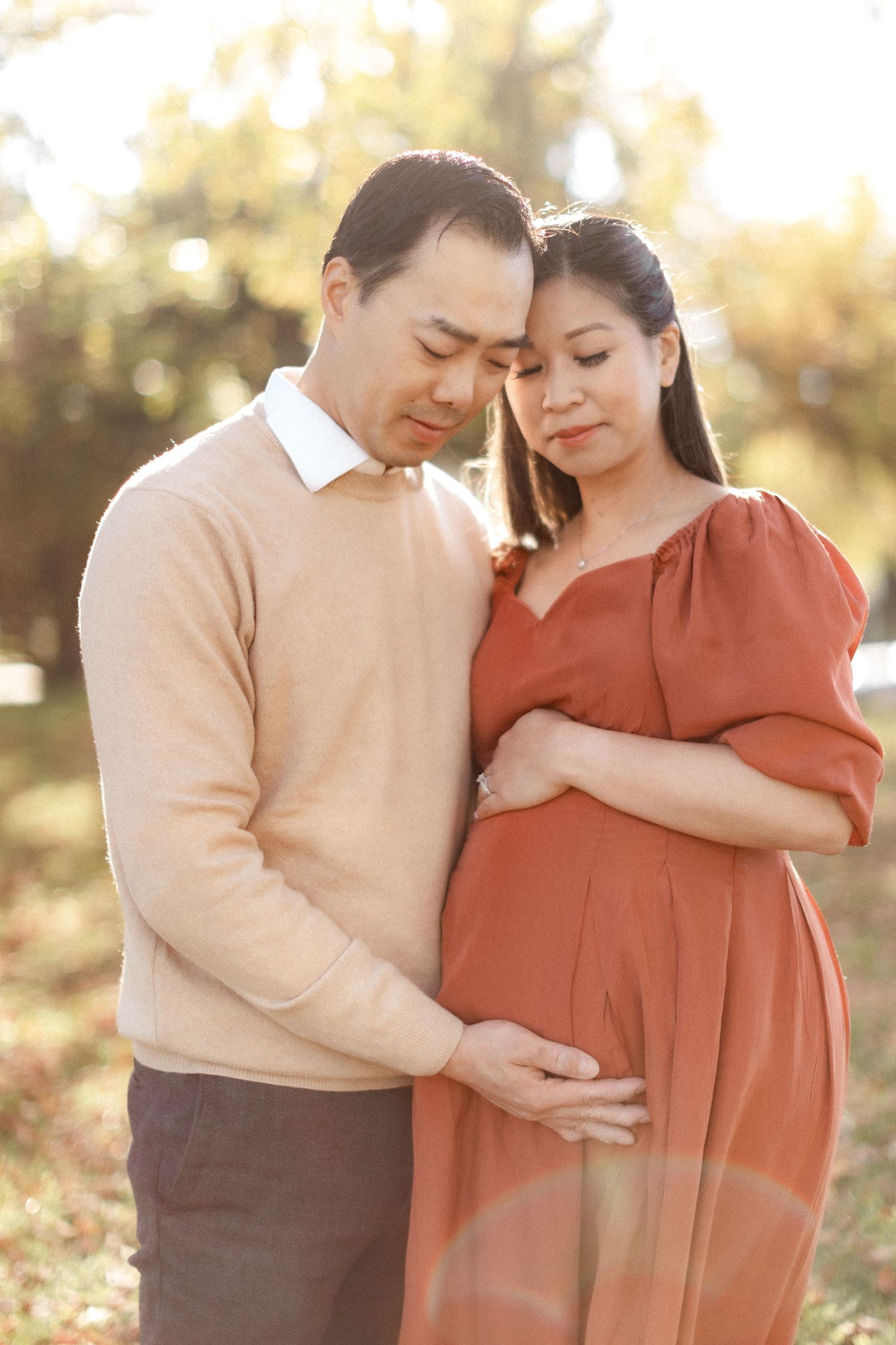 Intimate maternity photoshoot of couple in autumn sunlight with woman in rust-colored dress and man in beige sweater.