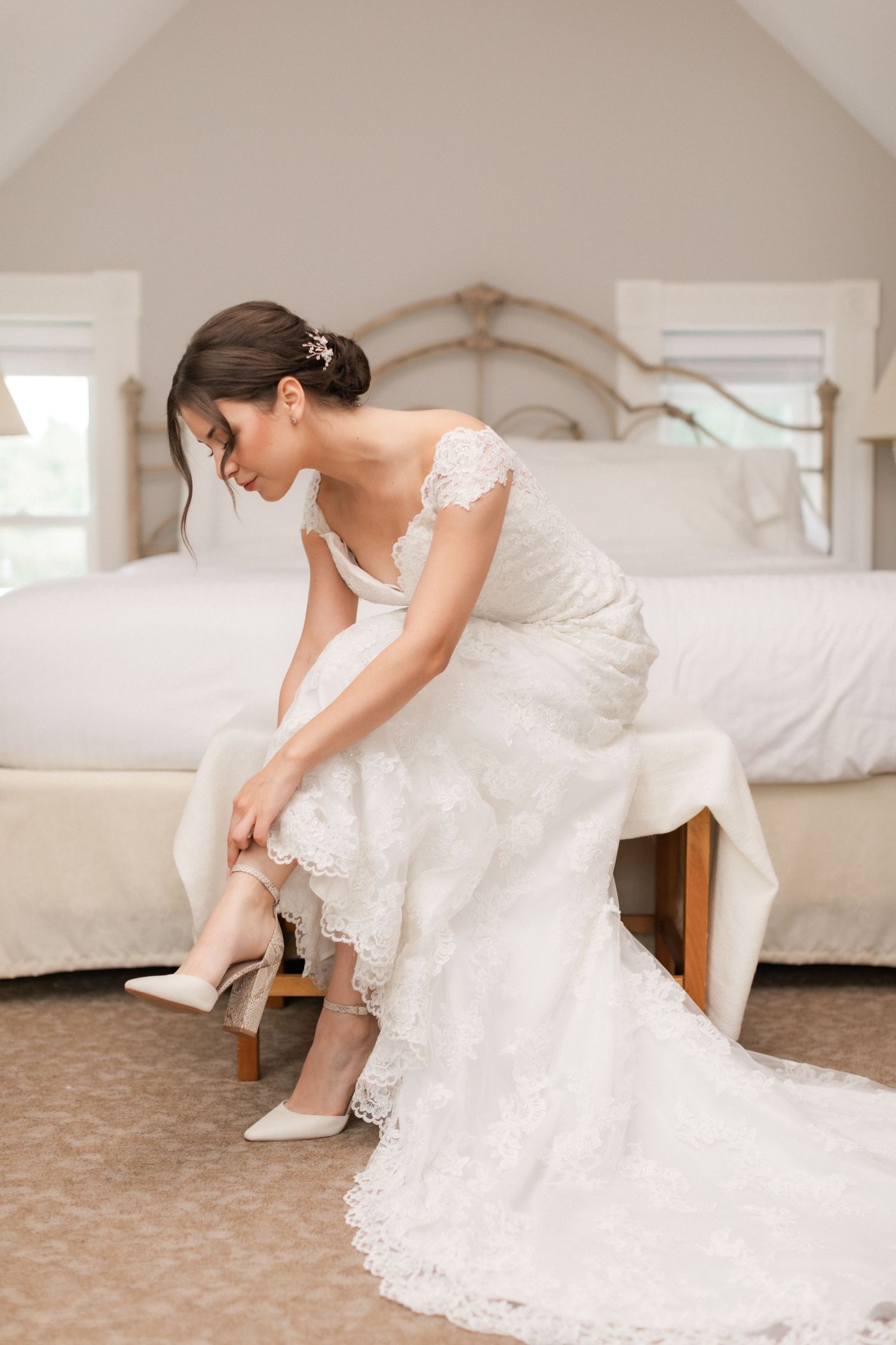 A person in a fitted lace wedding gown sits on a bed while putting on white heels.
