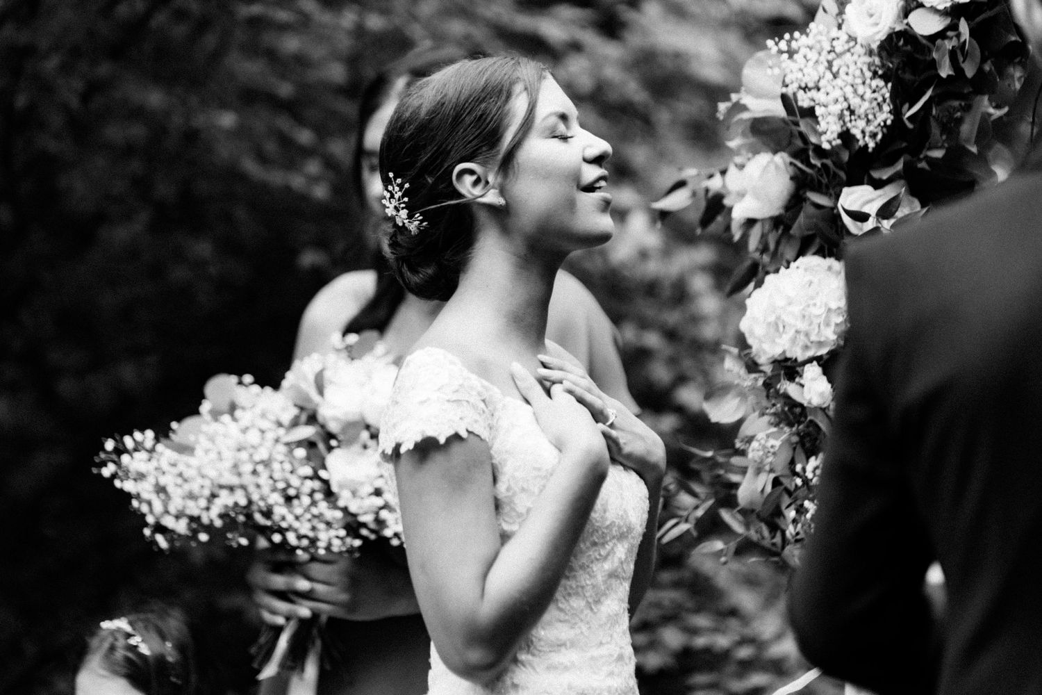 Black and white wedding photo showing bride with delicate floral headpiece in lace dress holding bouquet outdoors.