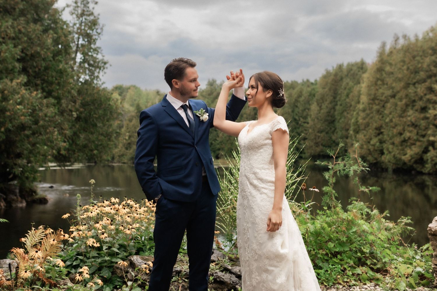 A couple shares a playful moment by a lake surrounded by trees with the bride in white and groom in navy.