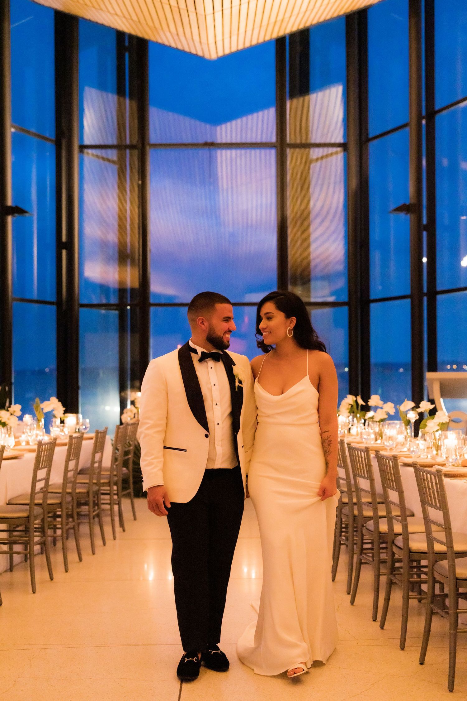 Couple standing in elegant reception venue during sunset with floor-to-ceiling windows.