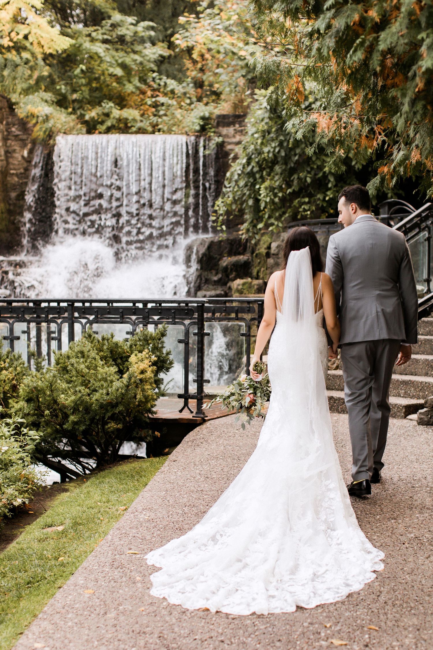 Wedding couple views waterfall from garden path with bride's lace train trailing behind.