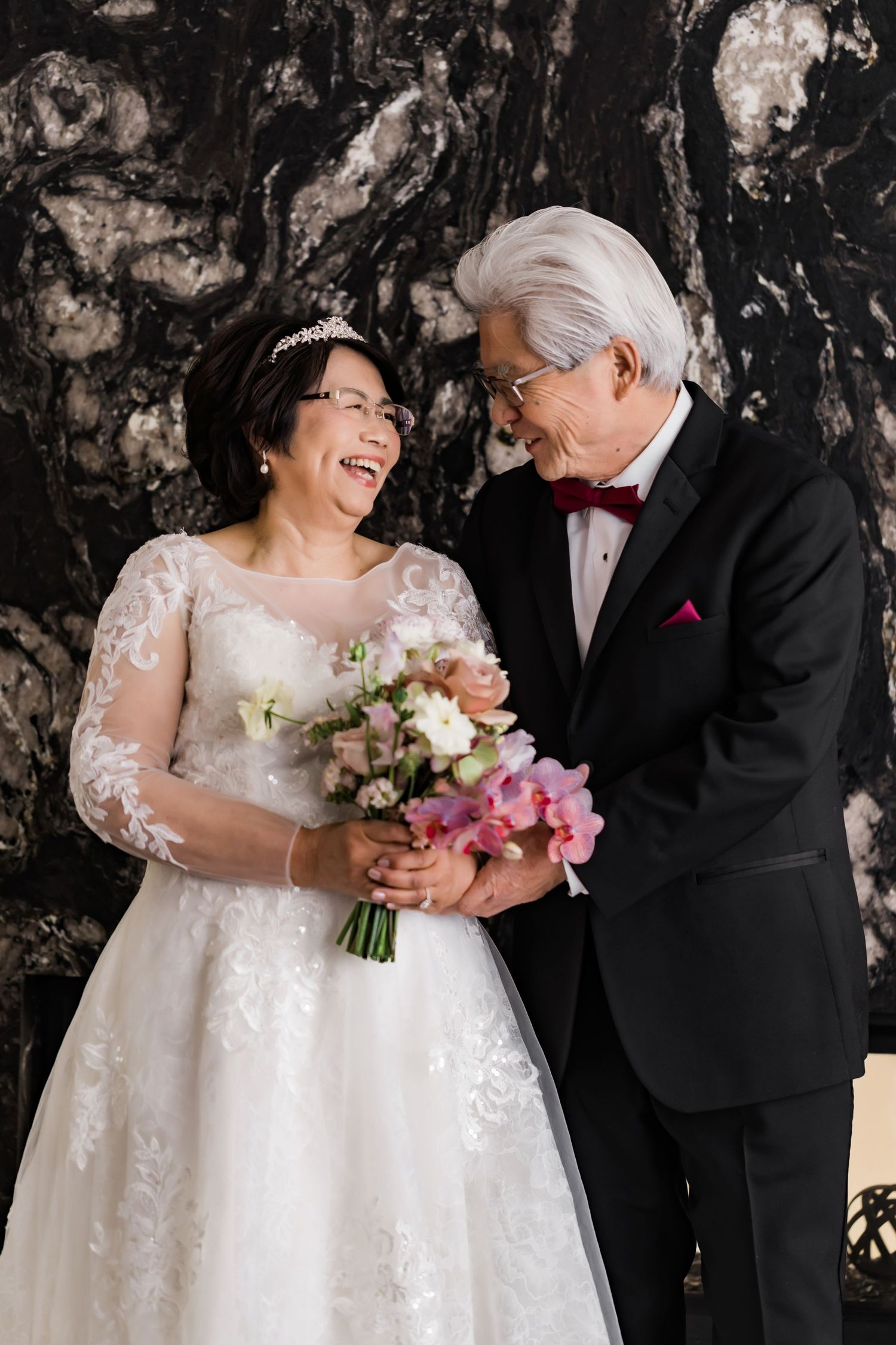 Heartwarming moment captured in sequence as couple shares joyful laughter during formal wedding portraits against dark stone wall.