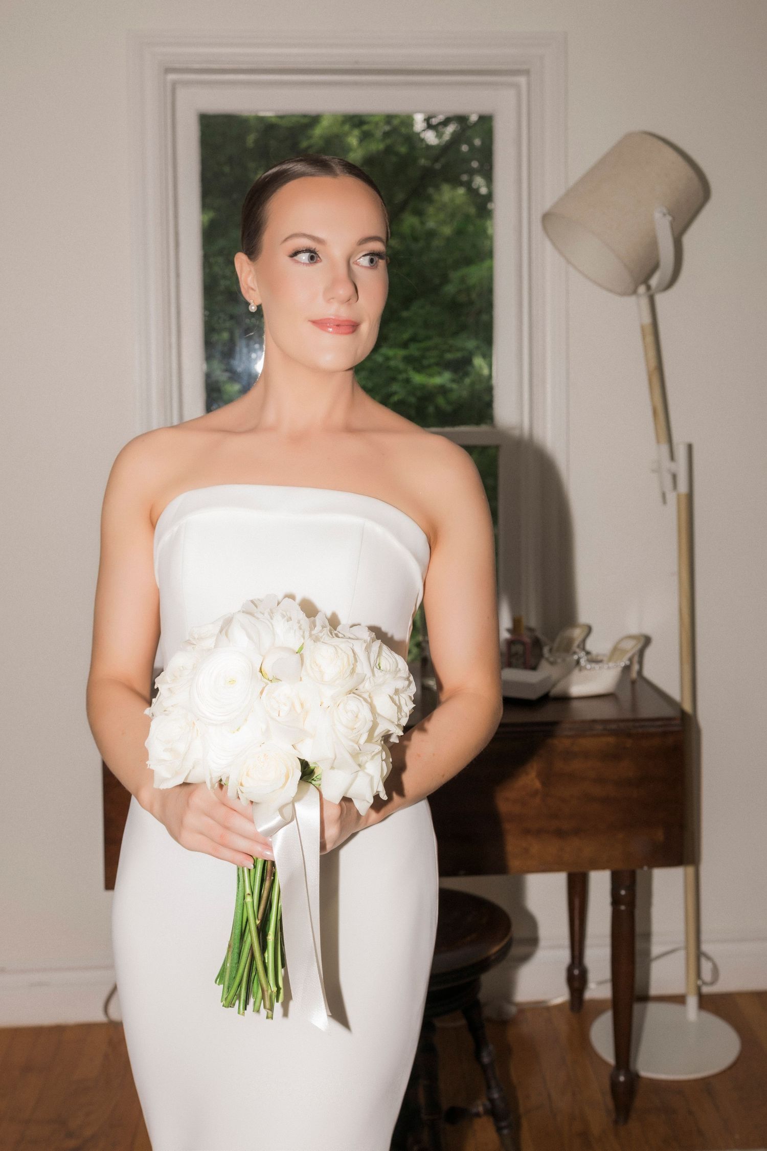 Bride in elegant white strapless dress holds a classic bouquet of white roses indoors.