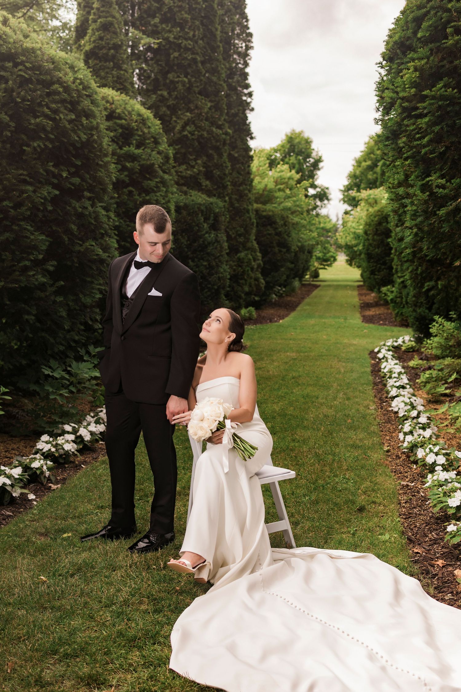 A wedding couple shares intimate moments along a garden path lined with evergreen trees and white flowers.