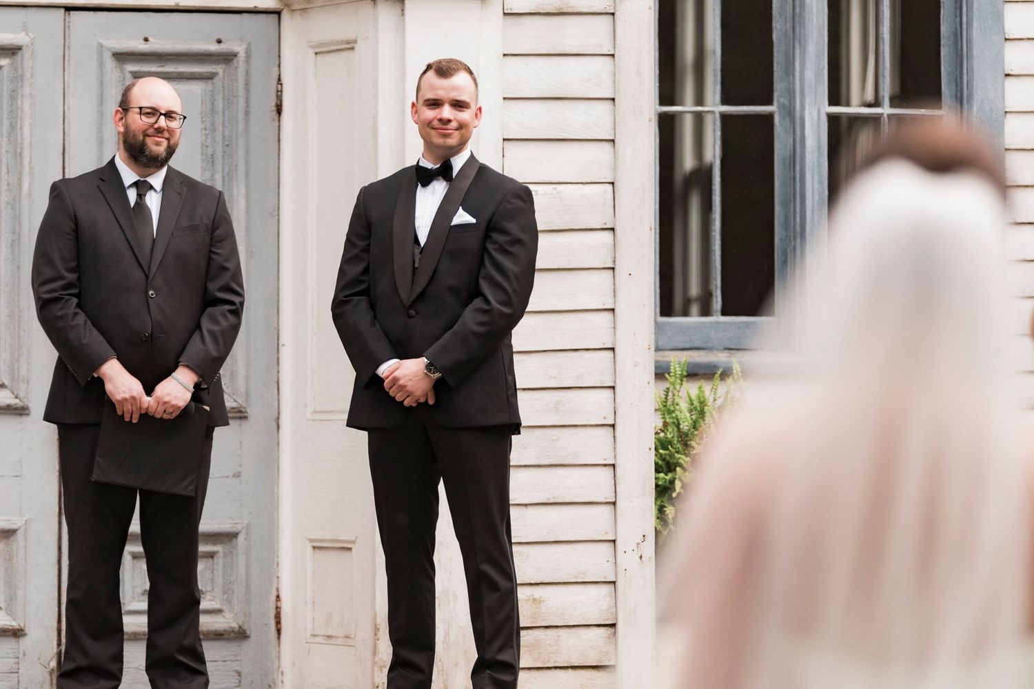Two men in black suits and bow ties wait expectantly as a blurred figure in white appears at the edge of the frame.