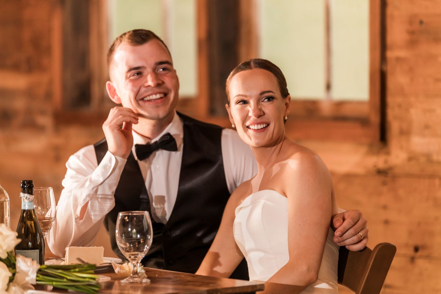 A couple shares a sweet moment while dining at a rustic reception with wooden walls and warm lighting.