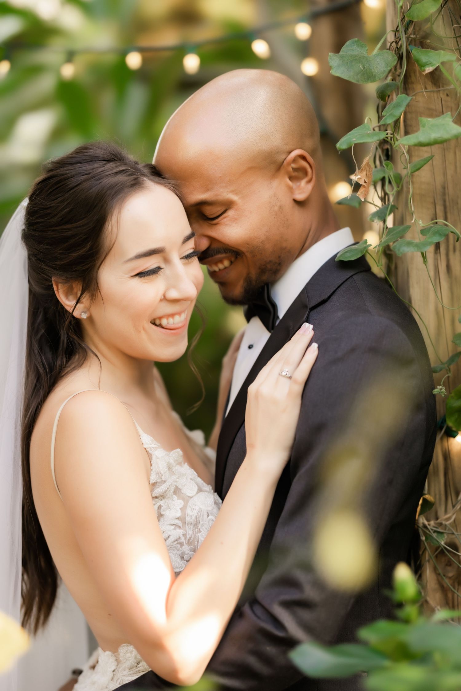 Close-up of bride and groom sharing tender moment surrounded by garden foliage and twinkle lights.