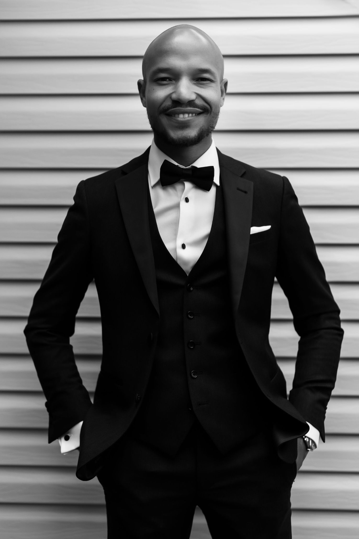 Black and white formal portrait showing classic three-piece tuxedo against textured wall.