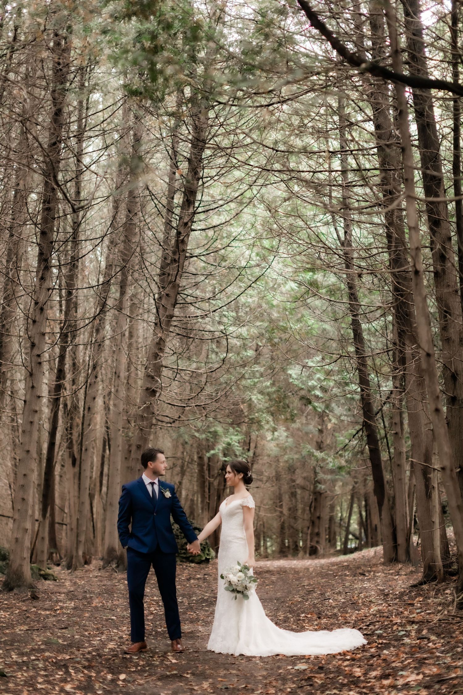 Bride and groom standing together on tree-lined forest path in romantic woodland setting.