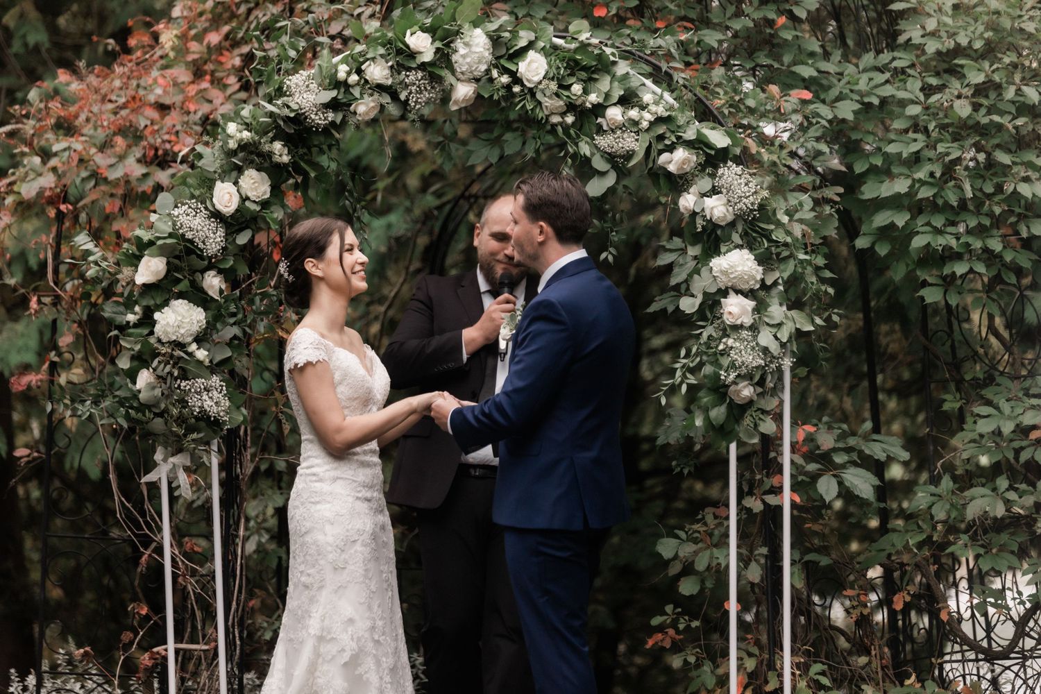 Intimate wedding ceremony moment under a white rose-covered arch in a garden.