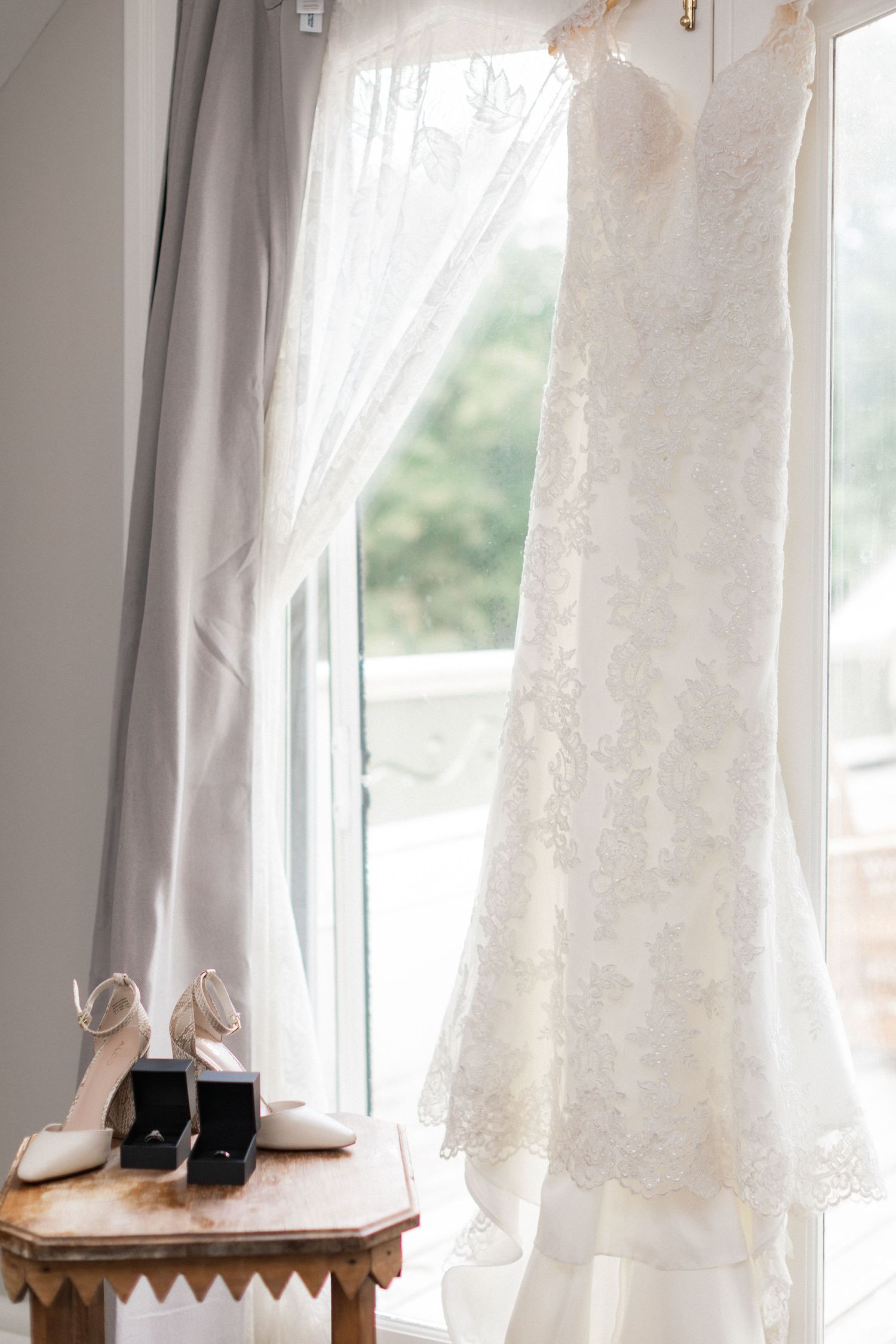 Elegant white lace wedding dress hanging by window with bridal shoes on side table.