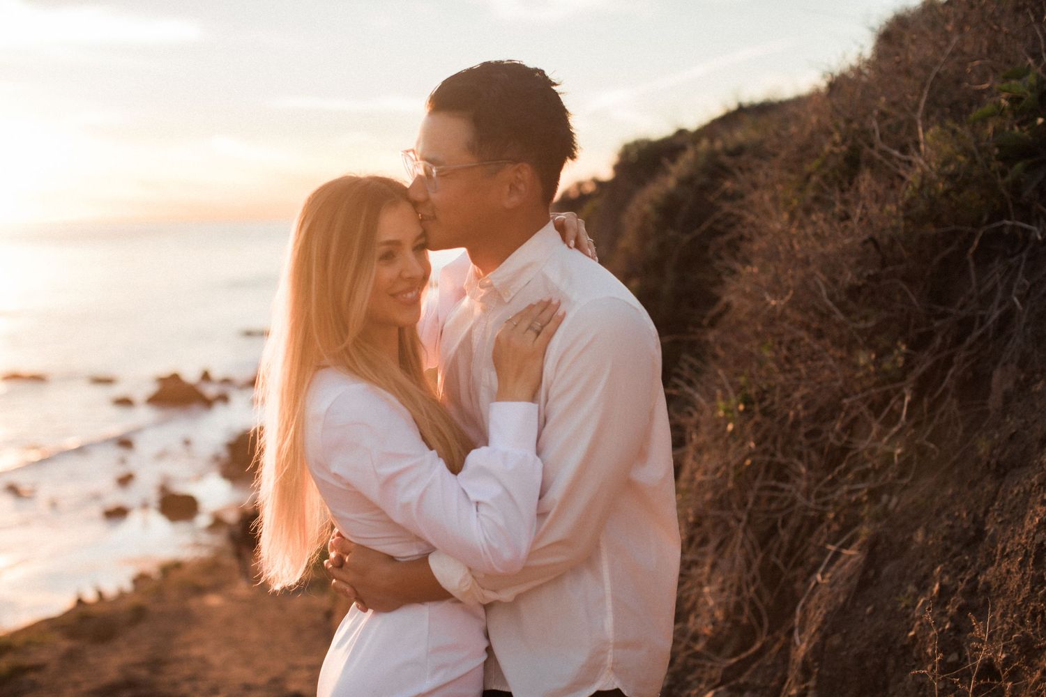 A tender moment between a couple in white clothing against a sunset coastal backdrop.