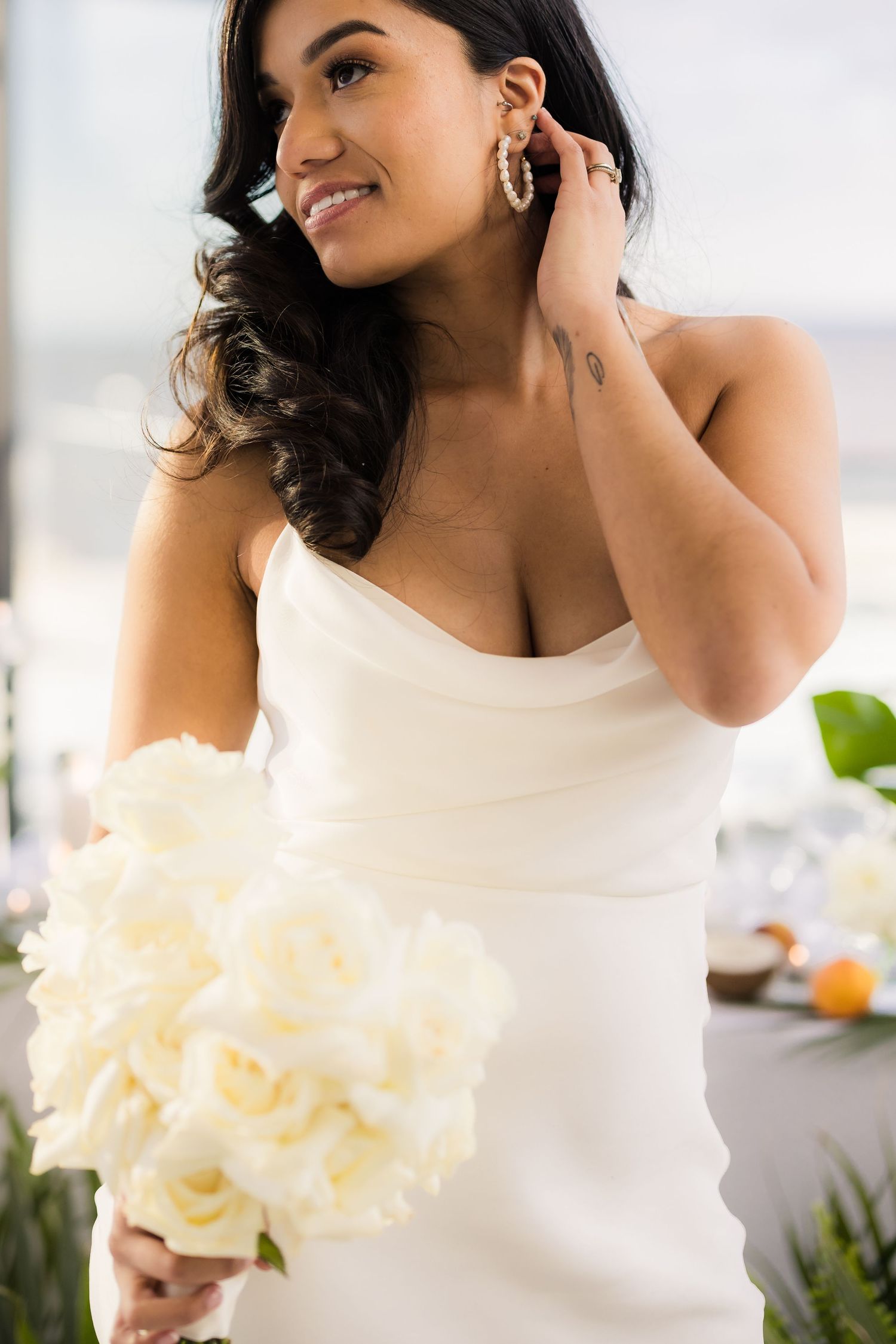Elegant bride in sleeveless white dress holding cream roses and wearing delicate hoop earrings.
