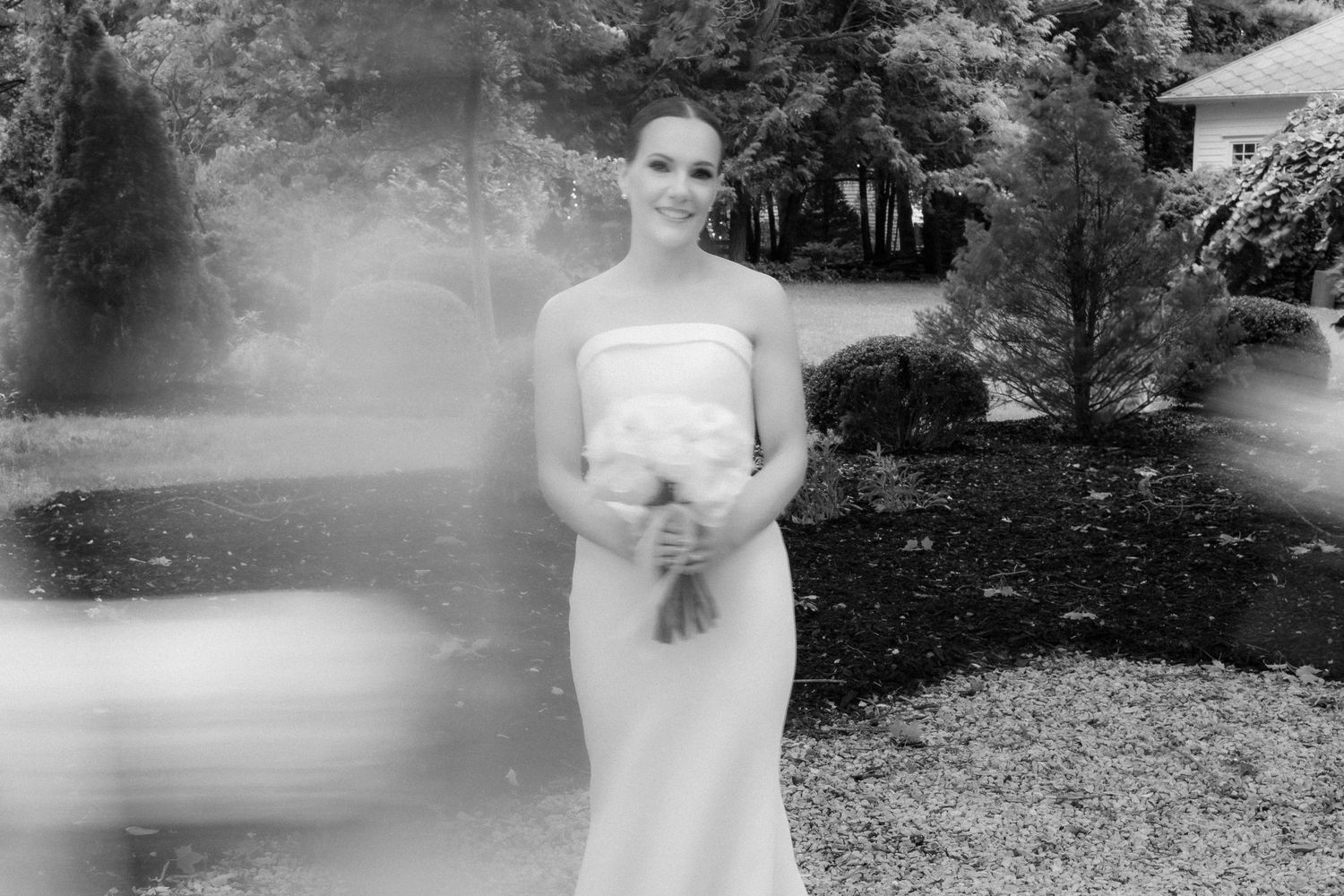 Black and white portrait of bride in fitted strapless dress holding bouquet in garden setting.