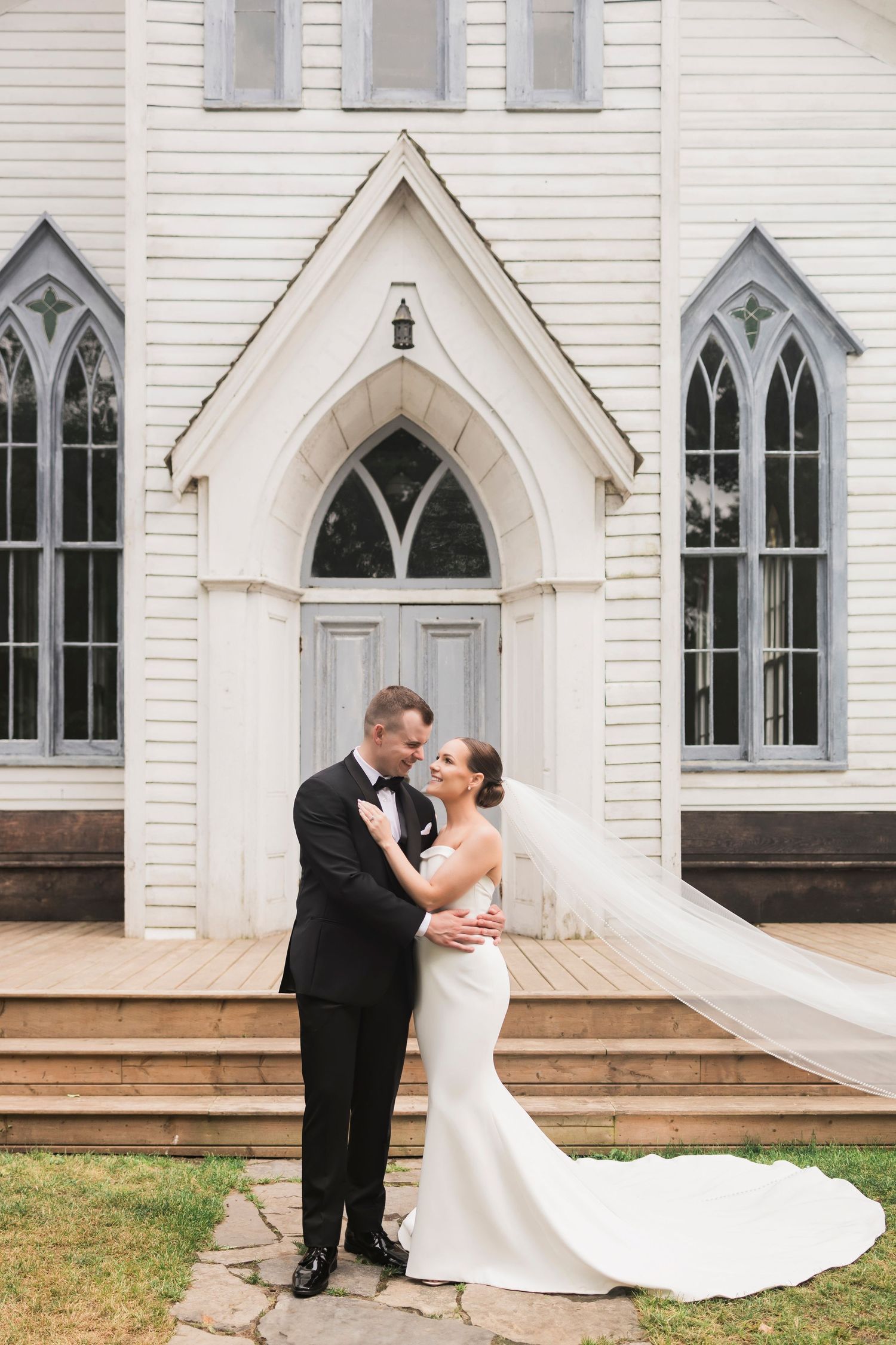 Elegant wedding couple poses in front of white gothic church with arched doorway and windows.