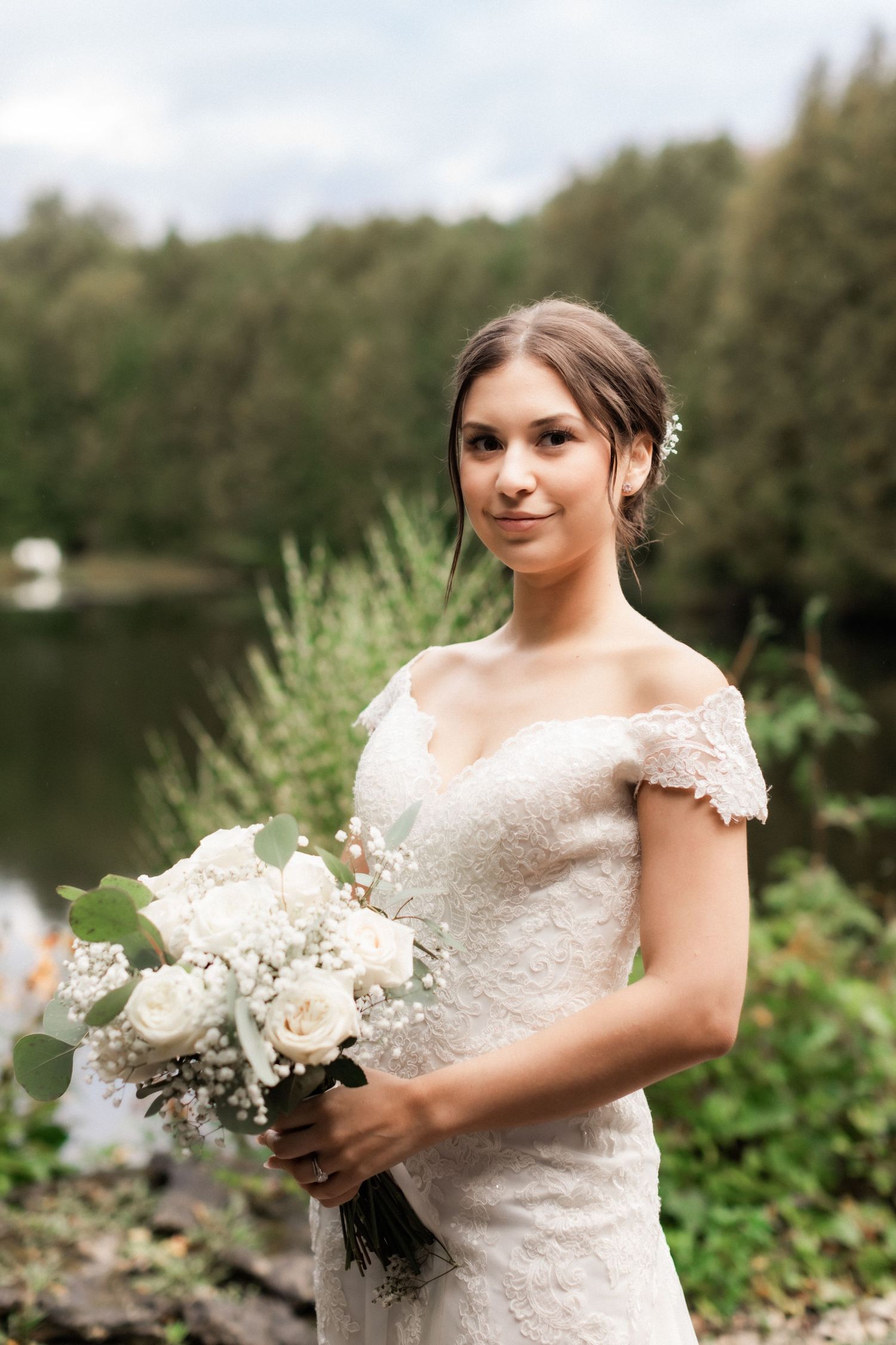 An elegant bride holds a white floral bouquet against a lakeside backdrop.