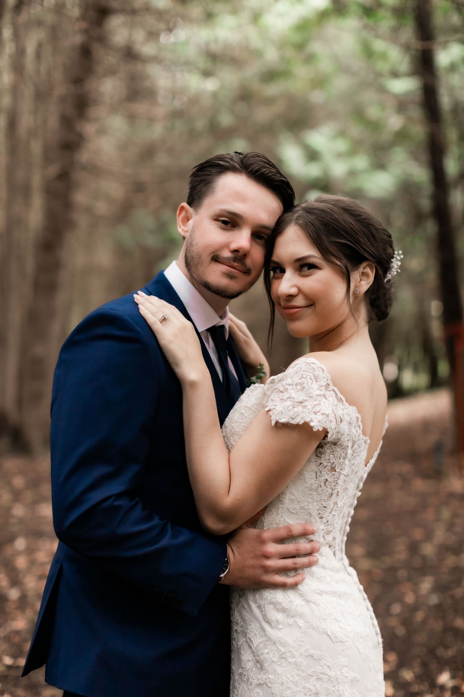 Elegant bride in lace gown and groom in navy suit pose intimately in forest setting.