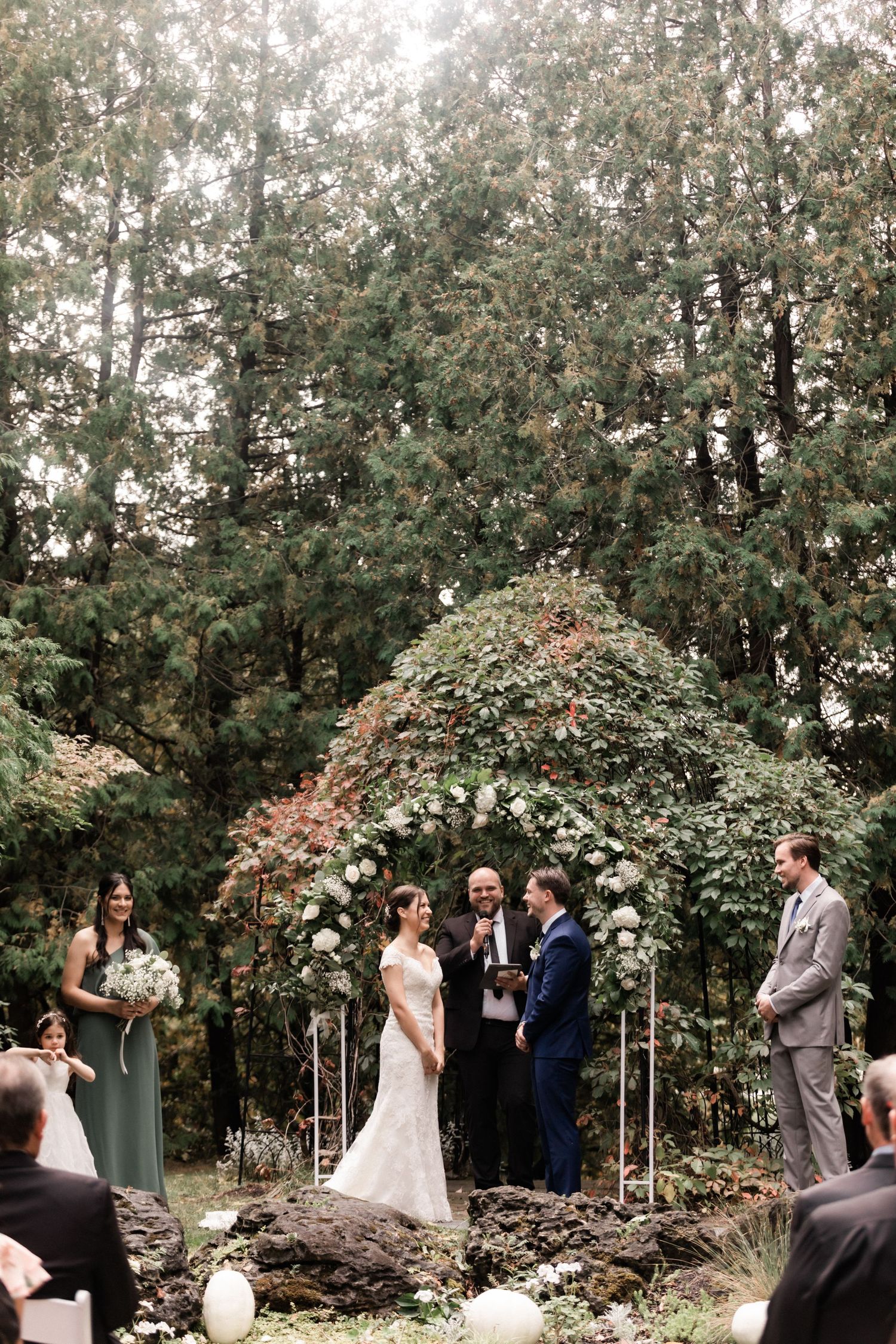Wedding ceremony under a floral arch in a wooded setting with white chairs and natural decor.