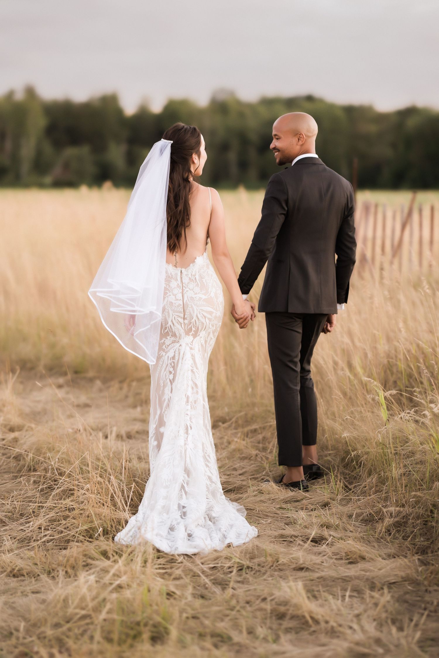 Back view of newlyweds walking through golden field, bride in intricate lace gown with veil.
