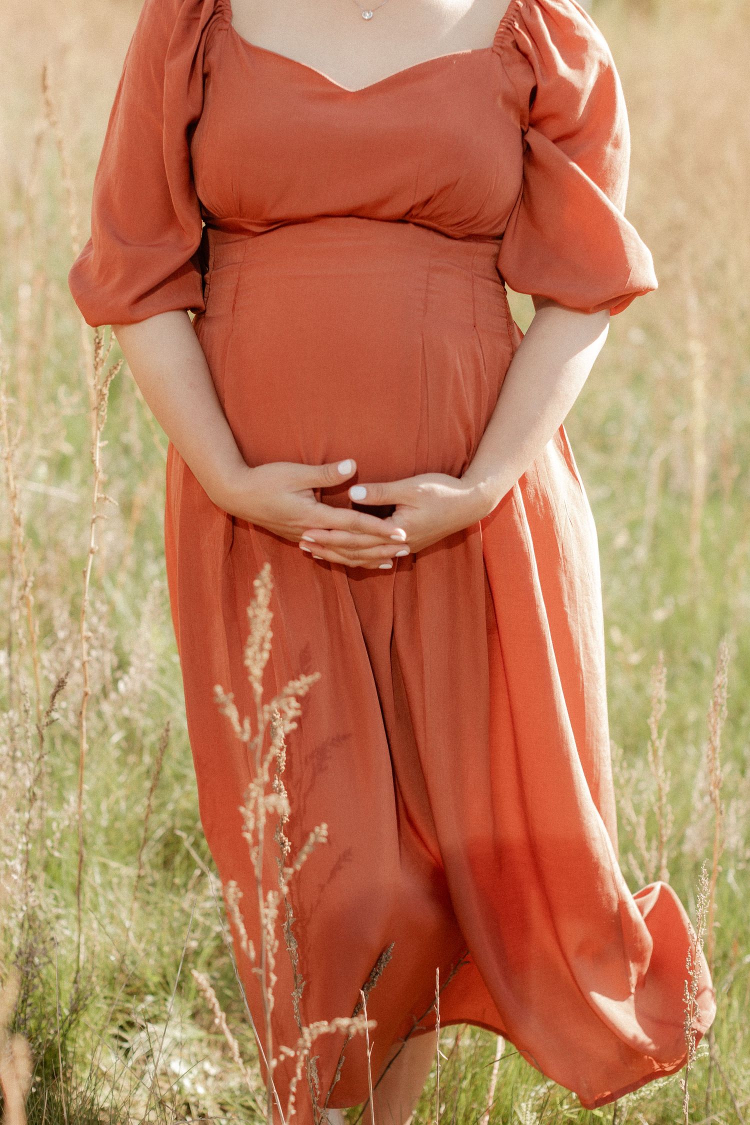A woman in a flowing rust-colored dress cradles her pregnant belly in a sunlit field.