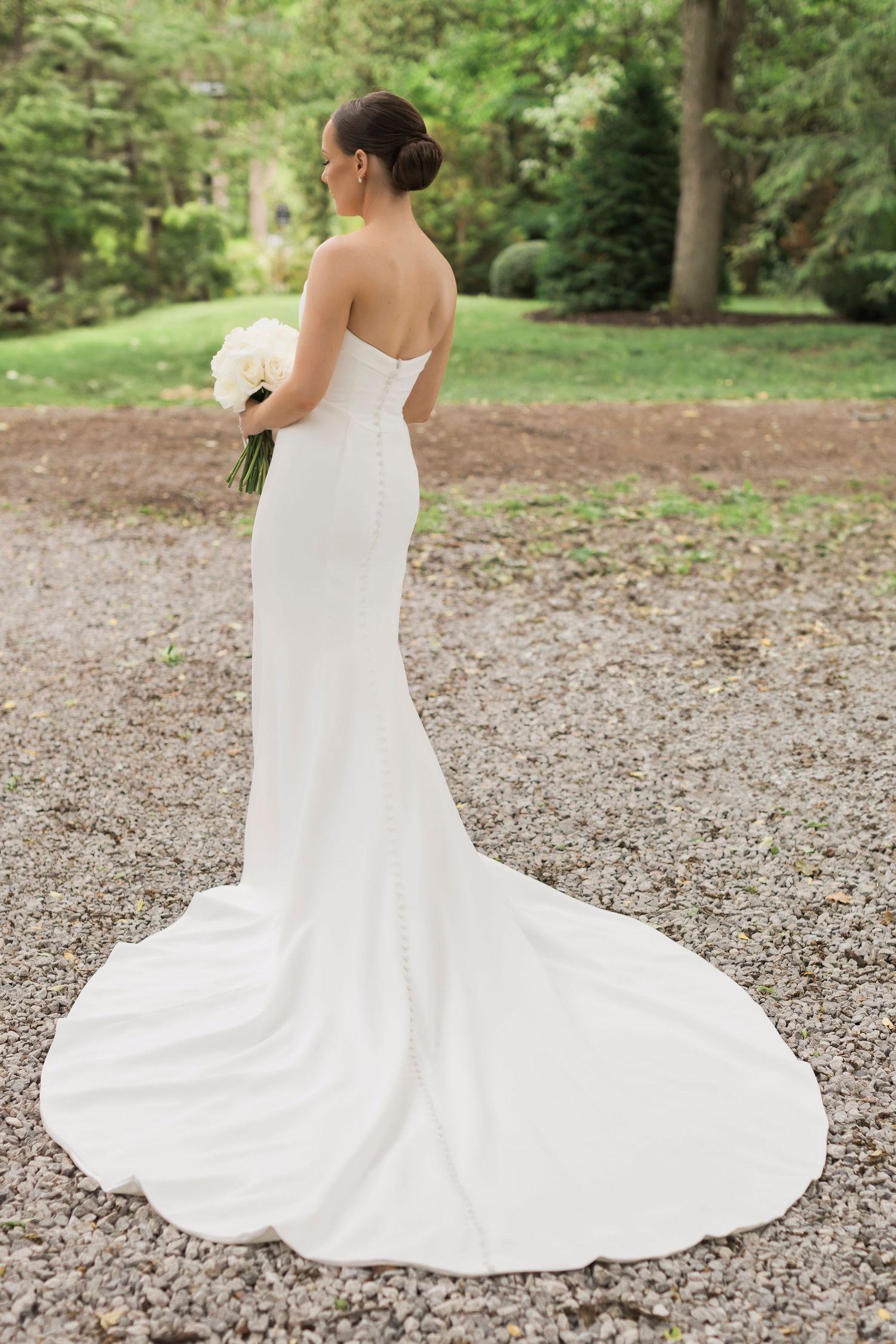 Back view of fitted strapless wedding dress with dramatic long train on gravel path surrounded by greenery.